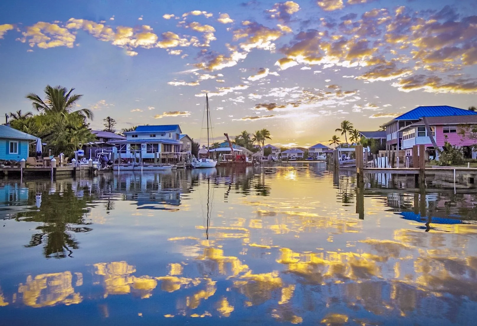 Colorful waterfront houses with docks and boats at sunset, palm trees, and a sky with scattered clouds reflected in the water.