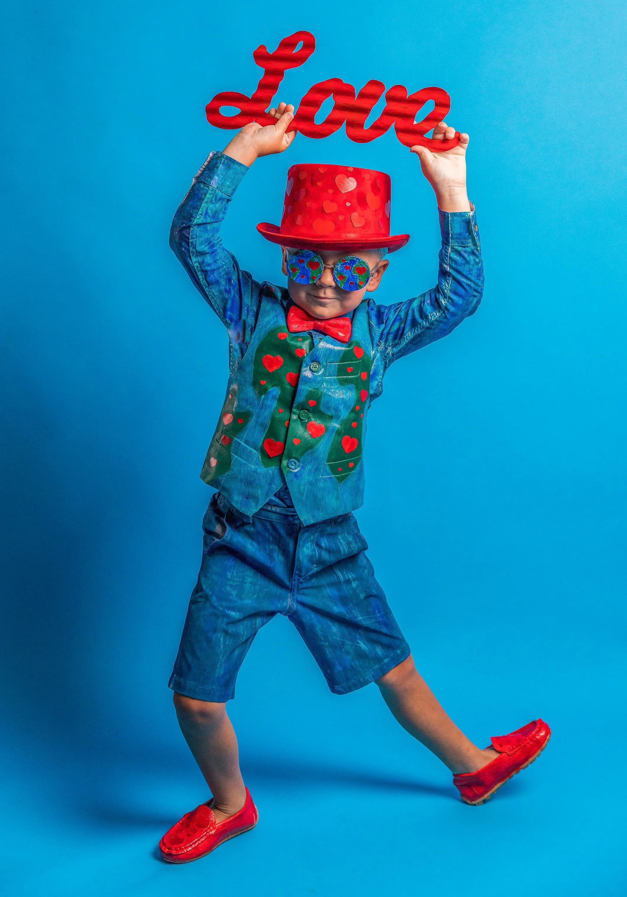 Child wearing red hat with hearts, colorful sunglasses, and red shoes, holding a red 'love' sign against a blue background.