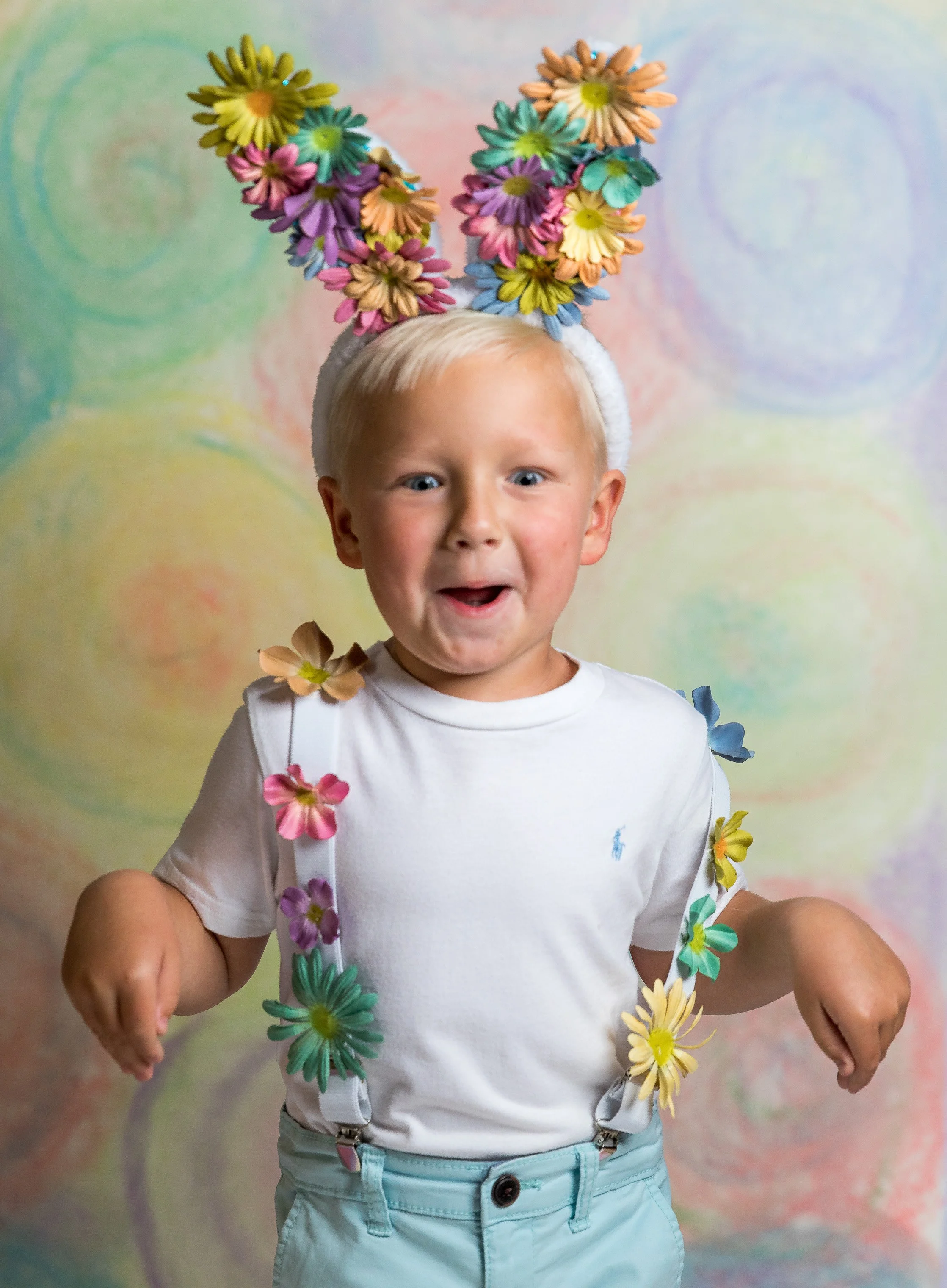 A young boy with blonde hair and blue eyes wearing a white t-shirt, mint green shorts, and suspenders. He is adorned with colorful artificial flowers on his head and arms, with a pastel swirl background.