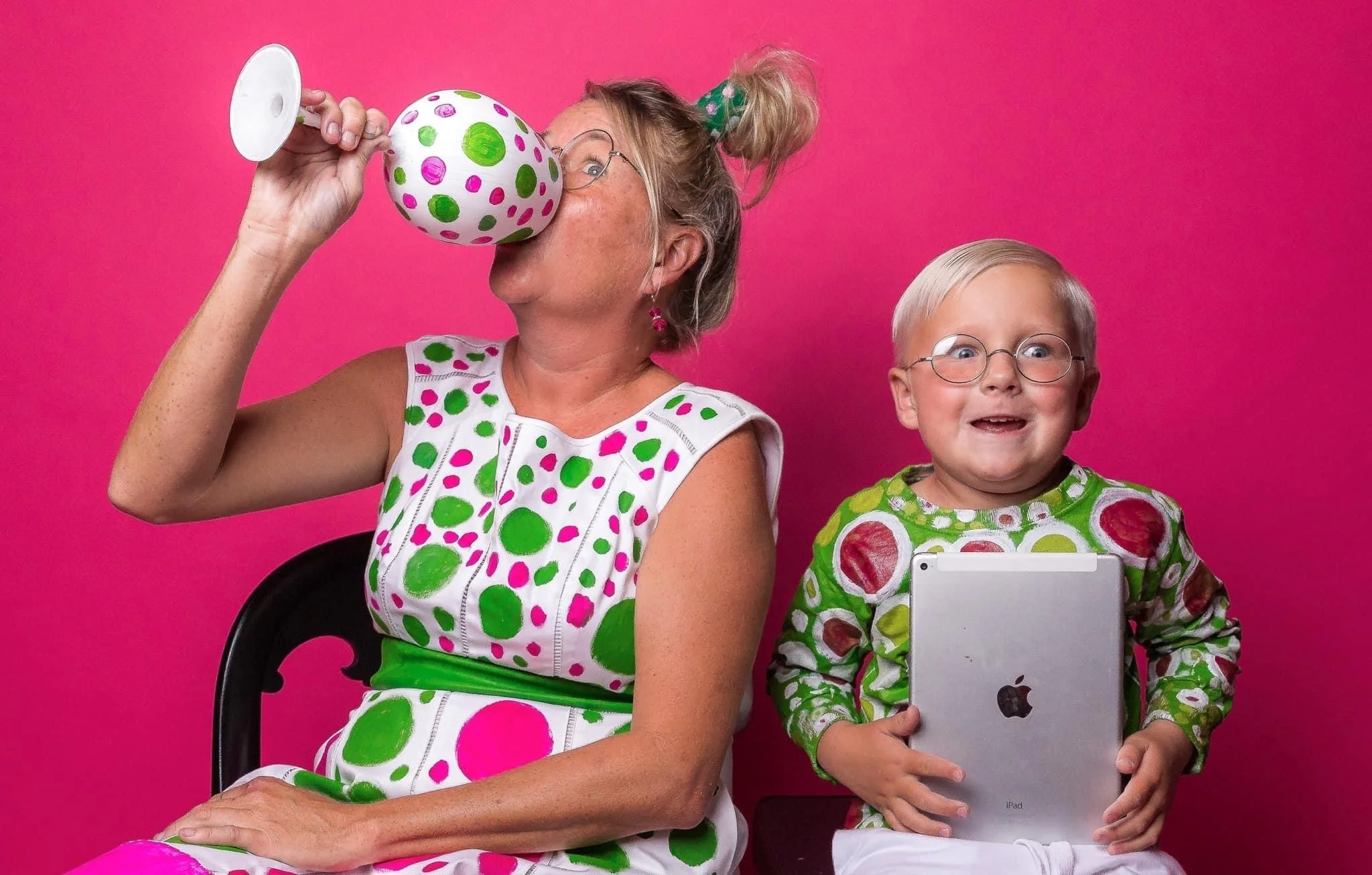 An older woman and a young boy, both wearing colorful polka-dot clothing, are sitting against a bright pink background. The woman is holding a decorated ball and appears to be looking through it, while the boy is smiling at the camera and holding a s