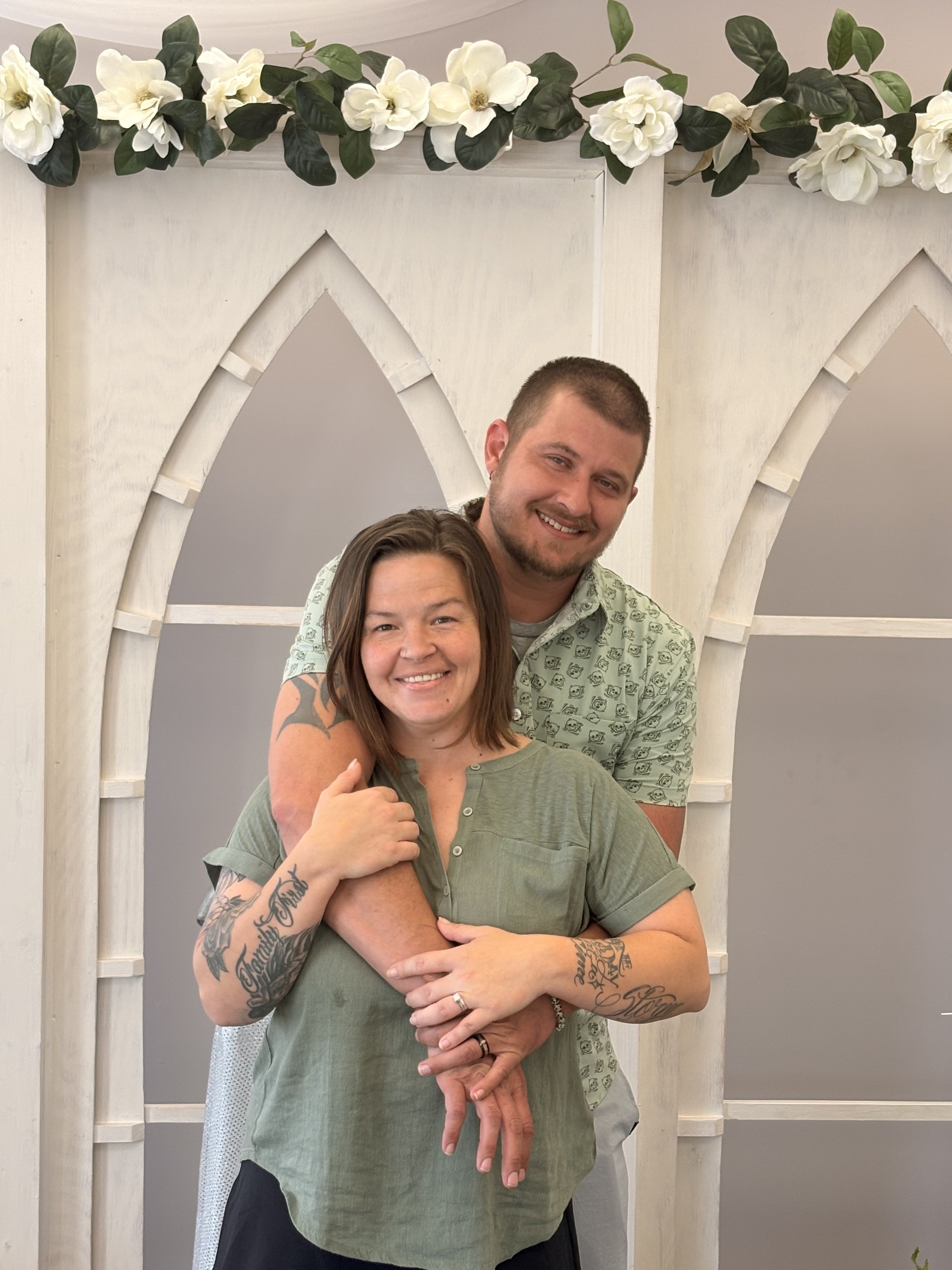 A smiling woman with tattoos on her arms and a ring on her finger stands in front of a man who is also smiling. He has short hair and is dressed in a patterned shirt. They are positioned against a backdrop featuring white wooden arches and a floral garland with white flowers and green leaves, suggesting a celebratory or special occasion.