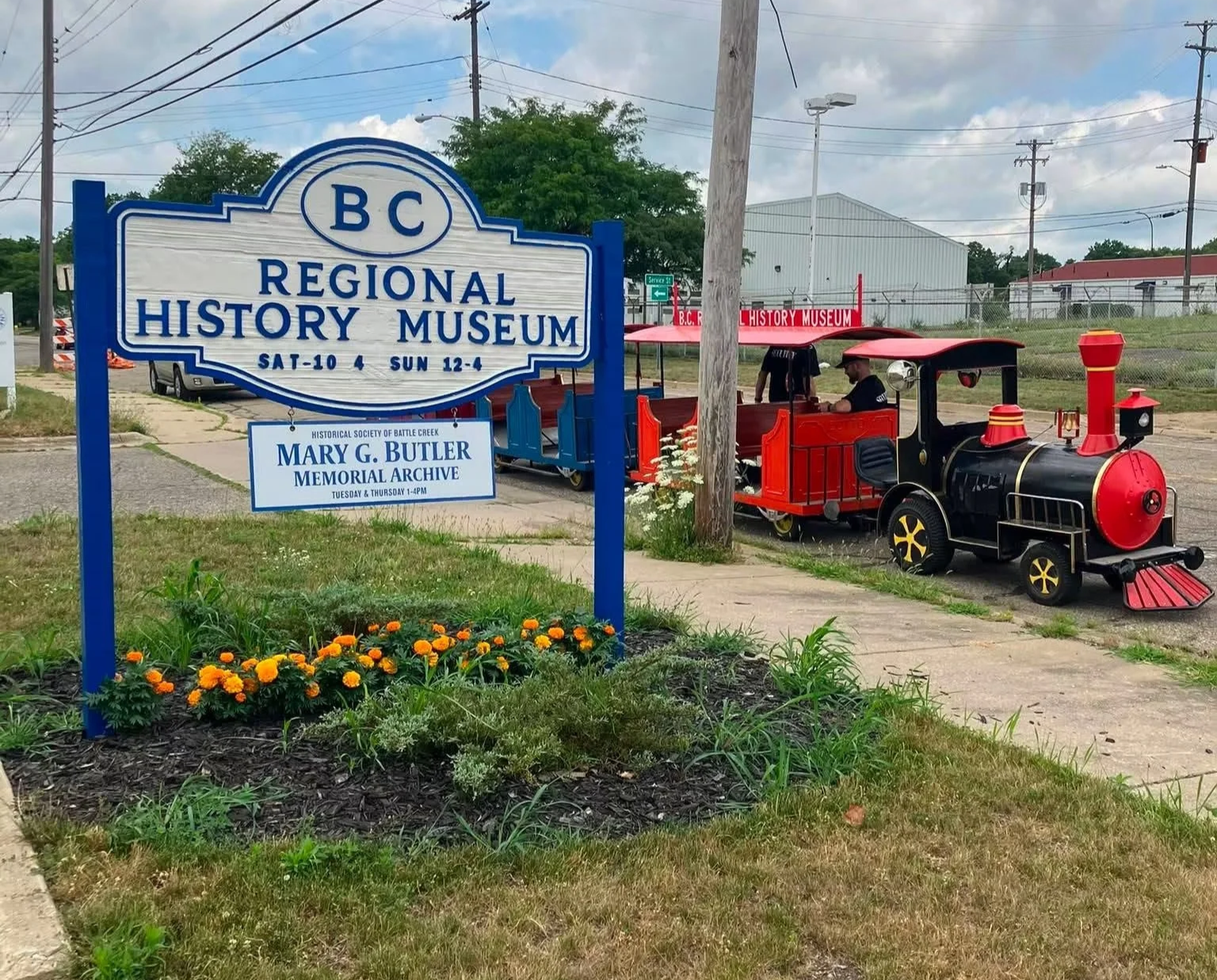 A sign for the BC Regional History Museum with information about visiting hours, and a small train replica nearby. The train has a black engine and red cars, situated on a paved area next to a sidewalk, with some flowers and grass in the foreground and a partly cloudy sky in the background.