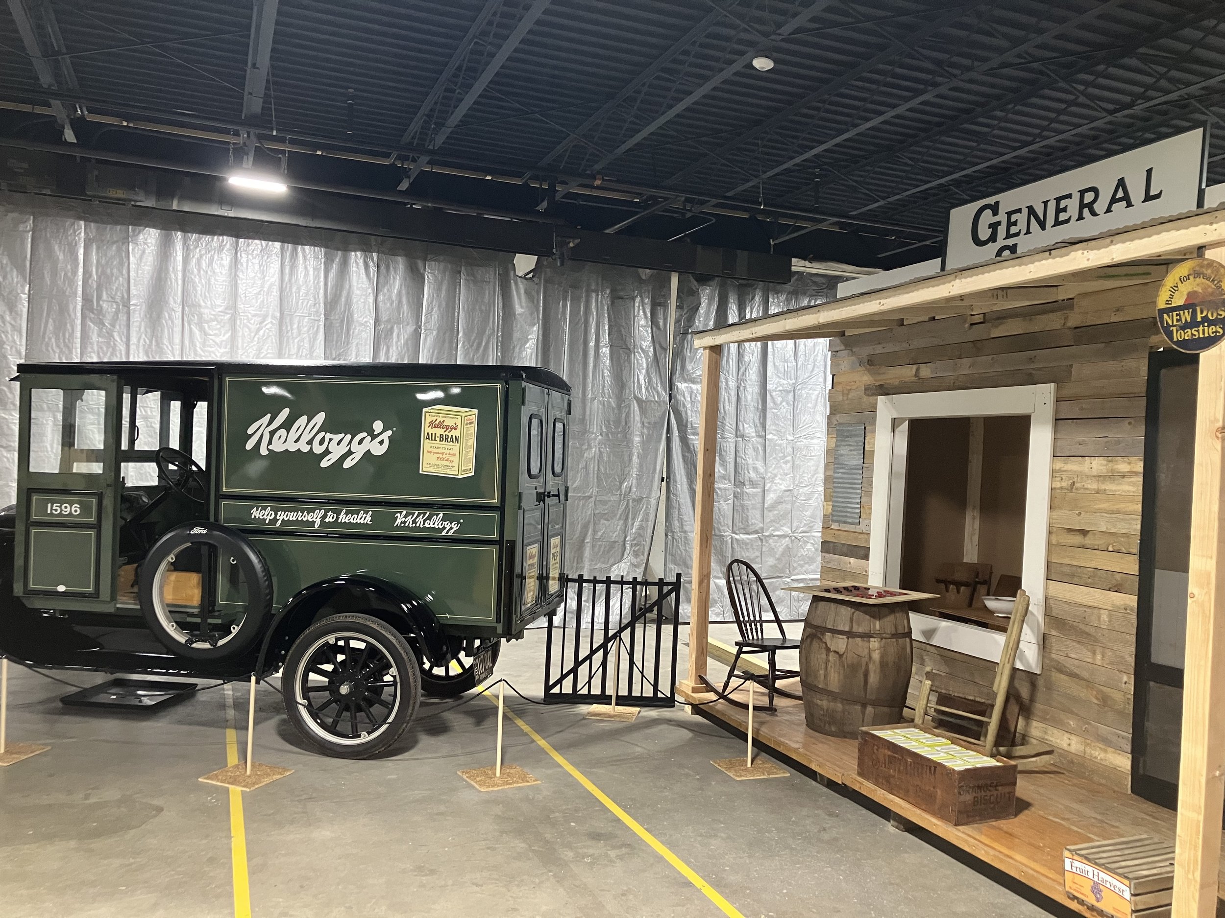 A green 1920's Ford delivery truck with white lettering reading "Kellogg's; Help yourself to Health. To the right of the truck is a covered porch on a wooden-board building. A checker game is set up on the porch.