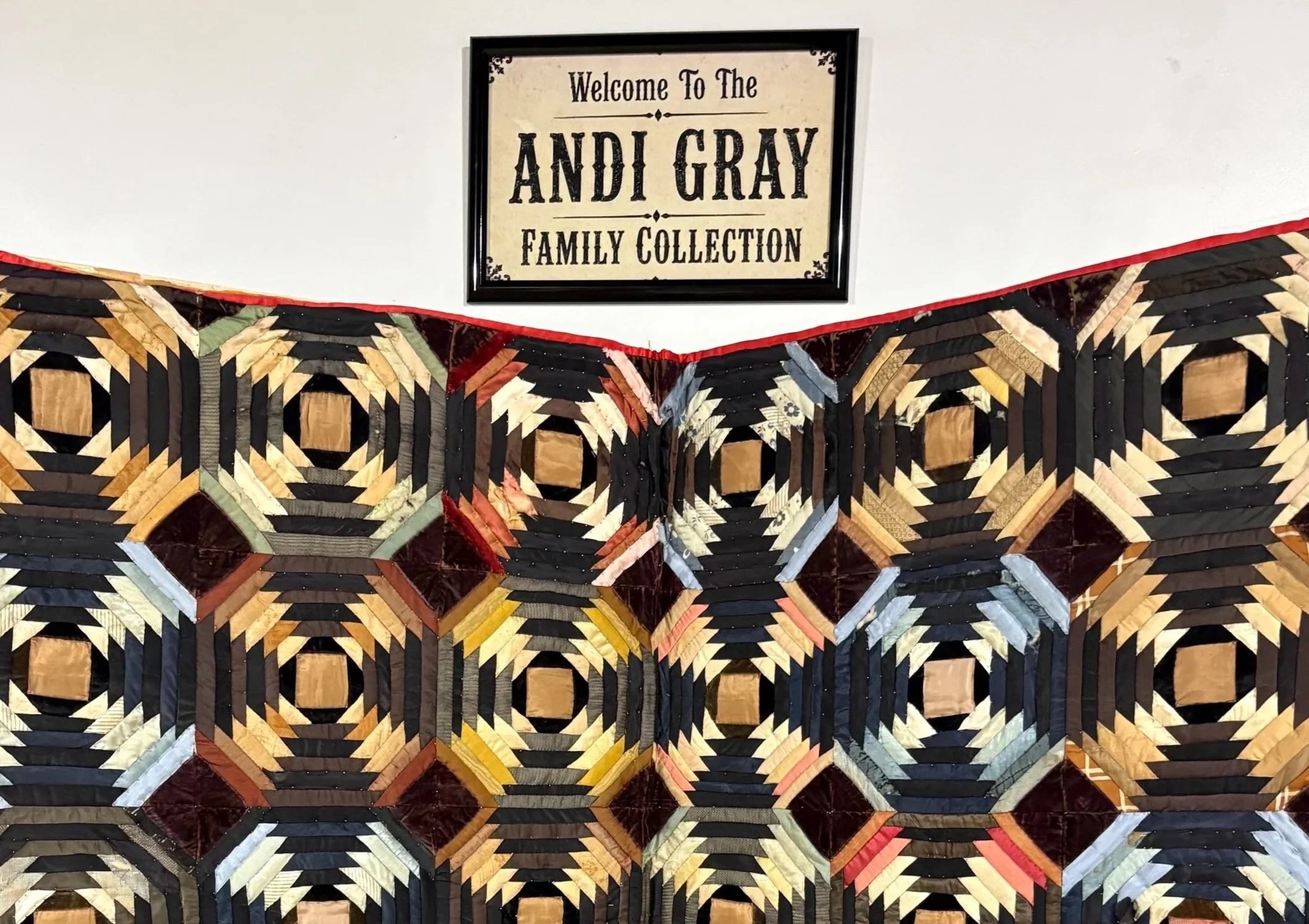 A sign hanging on a white wall which reads Welcome to the Andi Gray Family Collection, with a vintage quilt made of concentric circle blocks hung below it