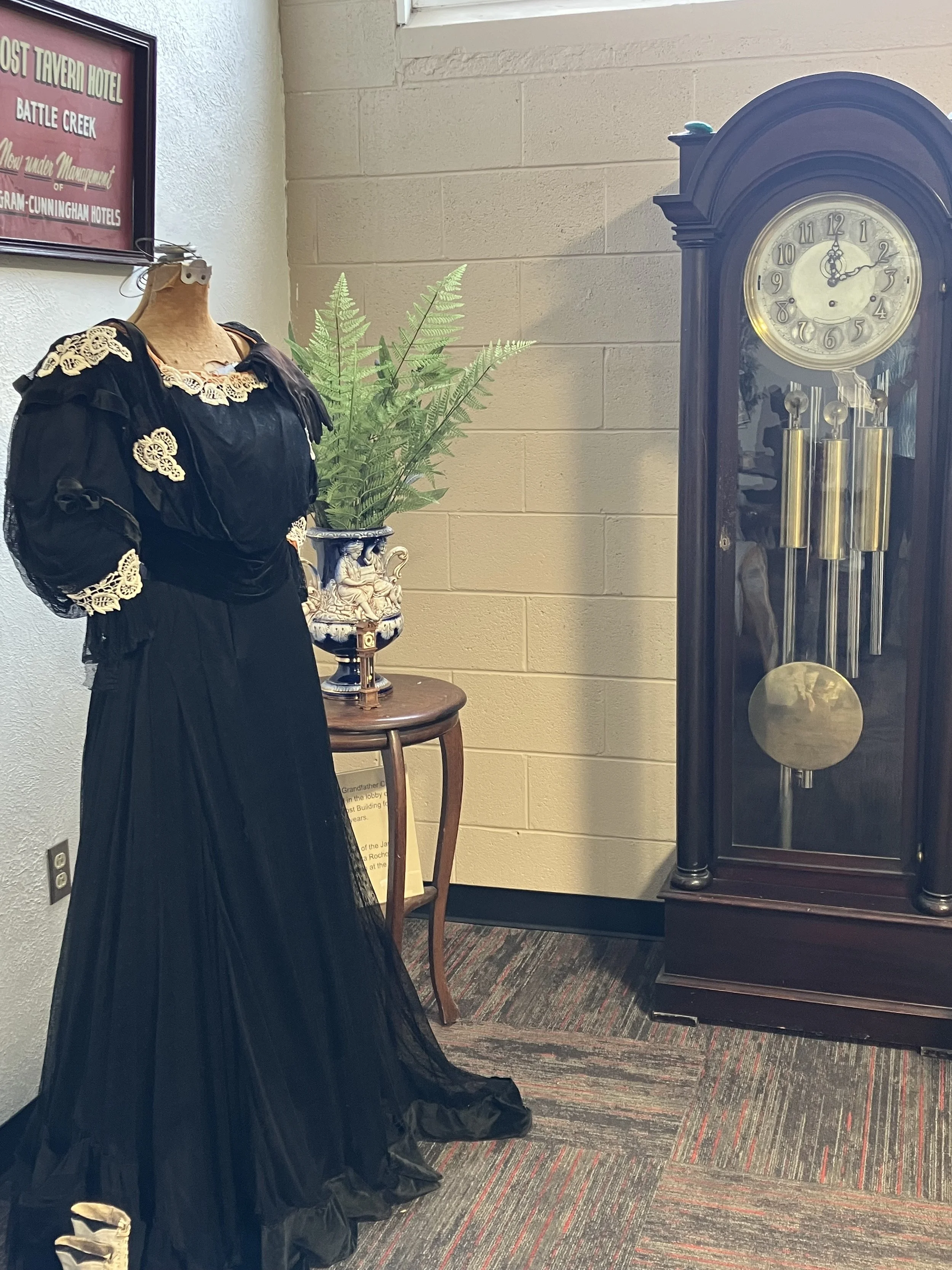 A clearly vintage black dress with long skirt and lace accents at the neck and shoulders is shown next to a fern in a fancy vase and a large grandfather clock.
