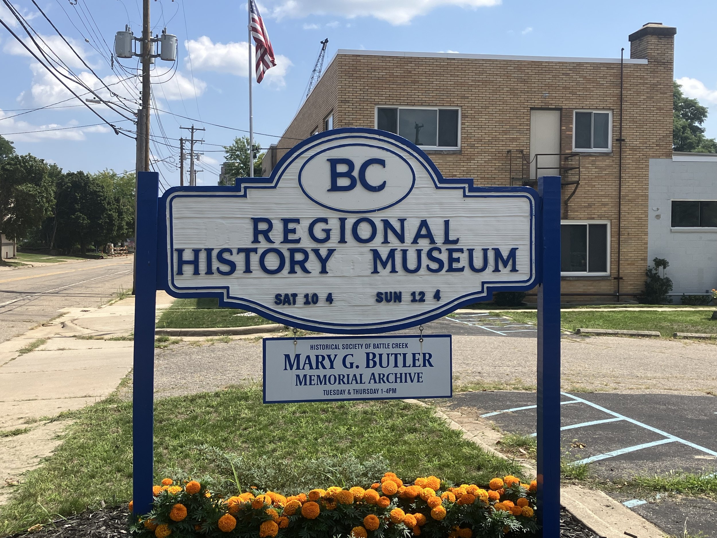 A blue and white sign saying Regional History Museum, listing the hours, with orange marigolds underneath