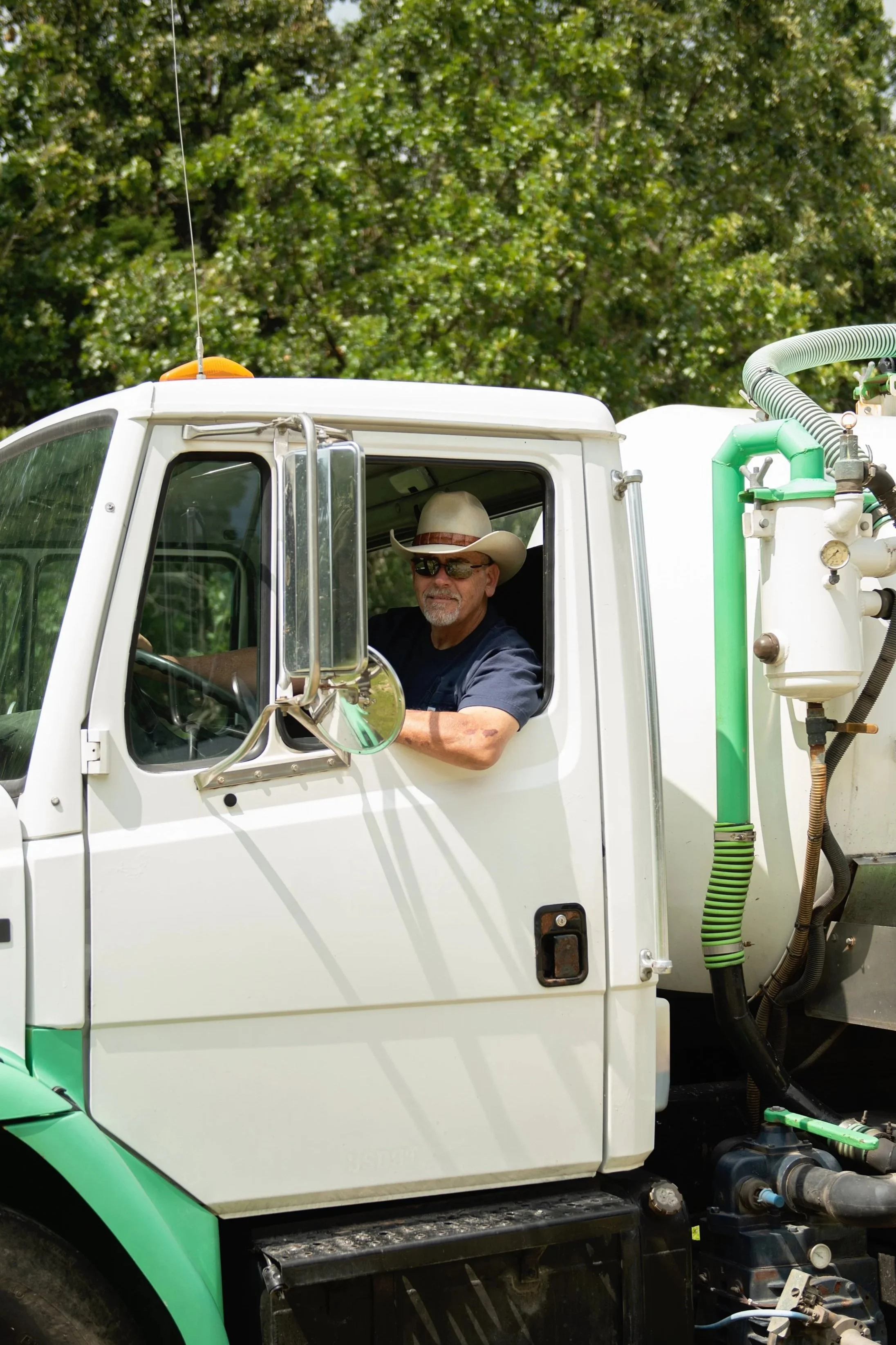 A man with sunglasses and a cowboy hat sitting in the driver’s seat of a white and green utility truck, with machinery and pipes on the side and a background of green trees.