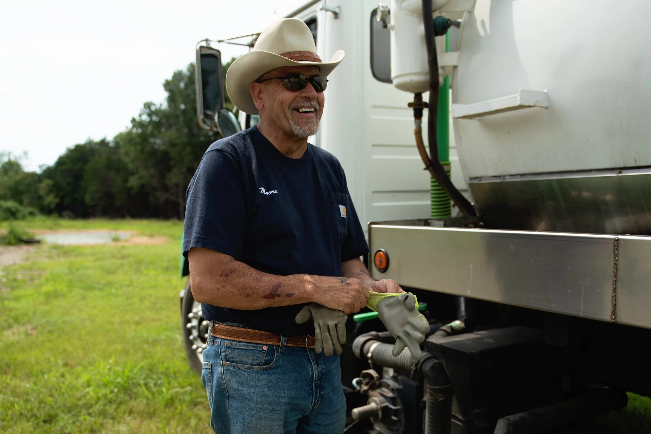 A smiling man wearing a cowboy hat, sunglasses, a dark shirt, and jeans is standing outdoors by a large piece of equipment, possibly a mobile kitchen or truck, holding a glove. There are trees and an open grassy area in the background.