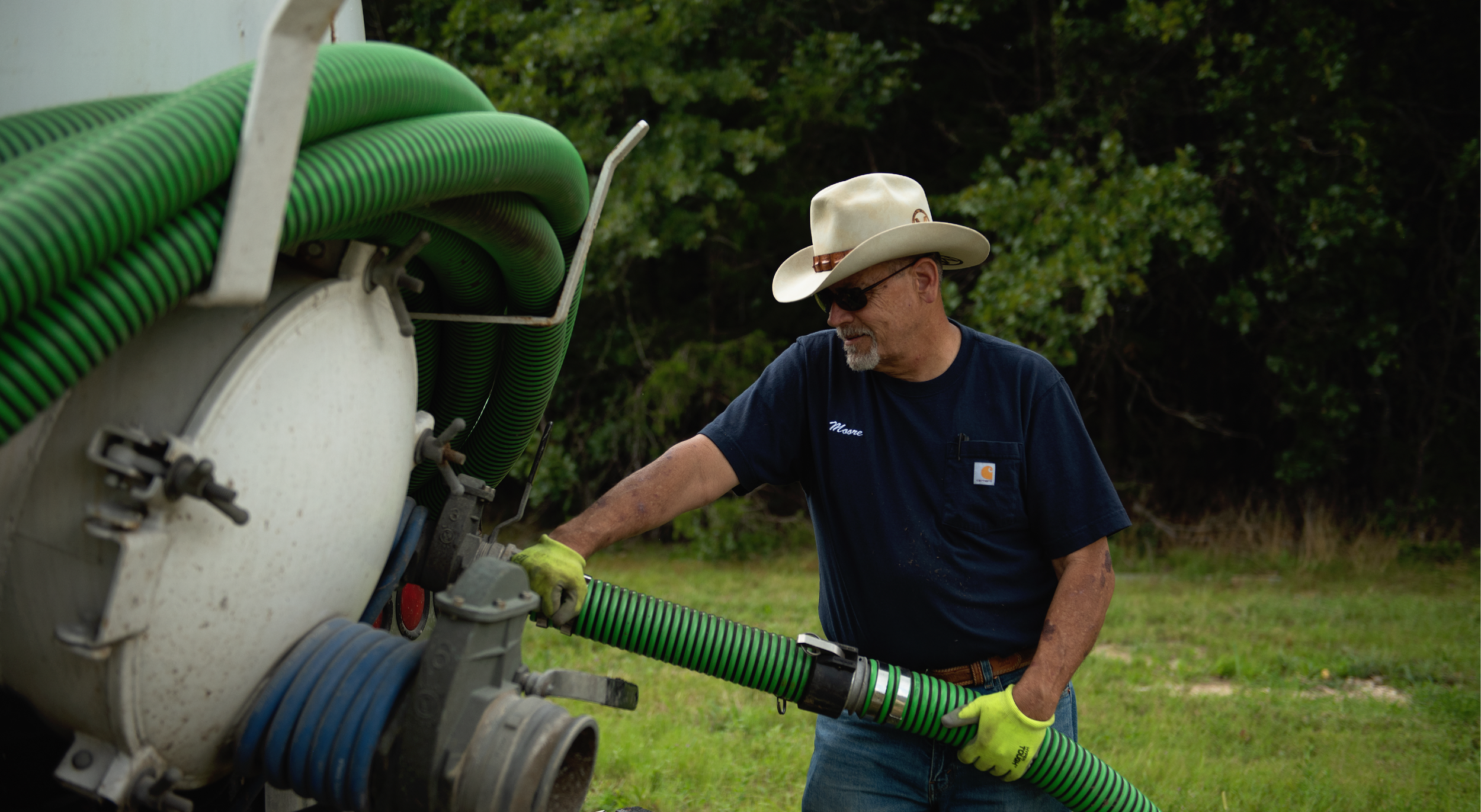 A man wearing a cowboy hat, sunglasses, and a navy blue t-shirt with a Carhartt logo, working outdoors with green hoses on a piece of equipment.