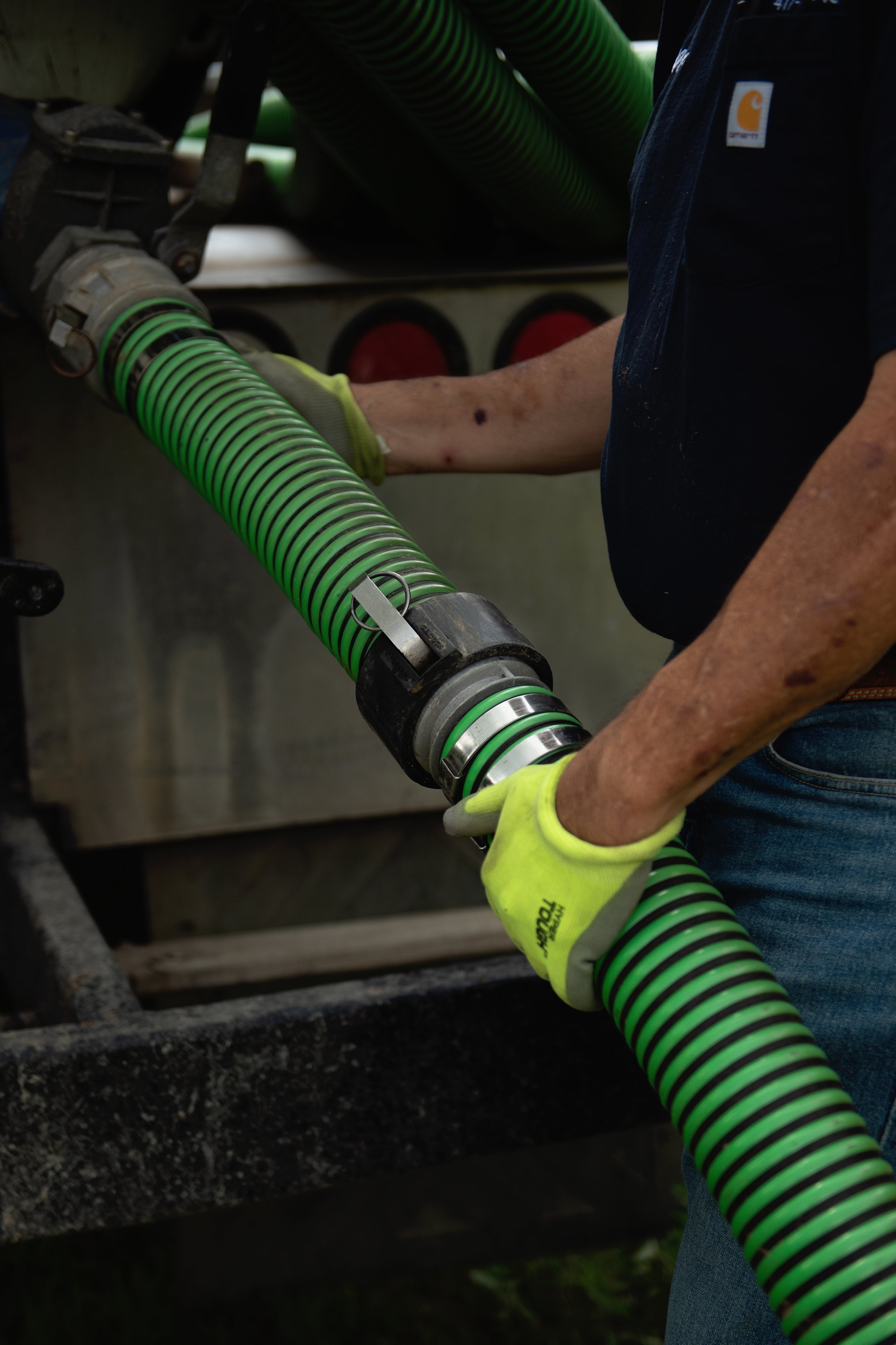 A person wearing a tan work glove and a dark blue Carhartt work shirt is handling a flexible green and black storm drain pipe near a utility truck, suggesting work related to construction or plumbing.