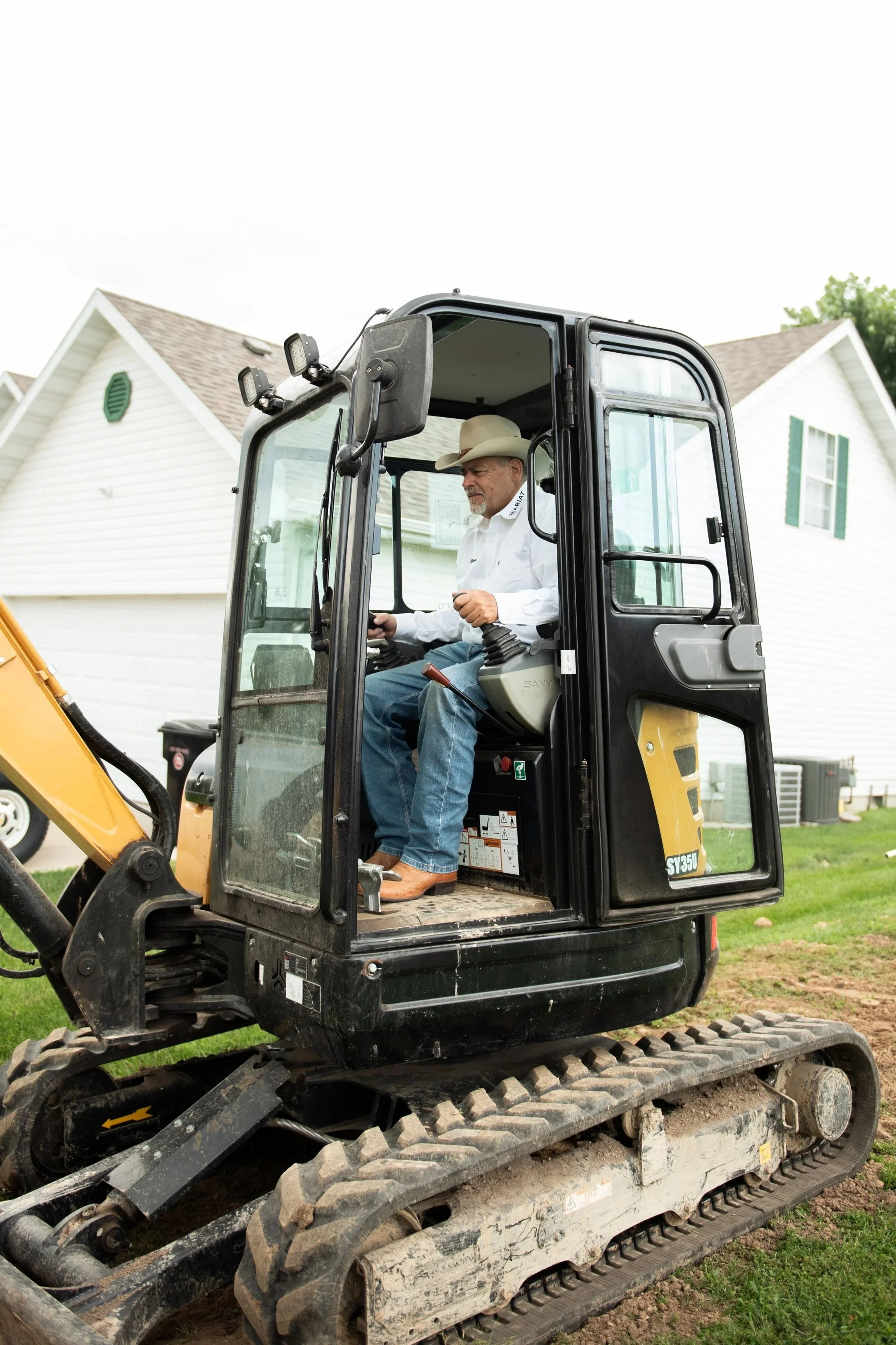 A man wearing a cowboy hat and white shirt operating a small excavator with a house in the background.