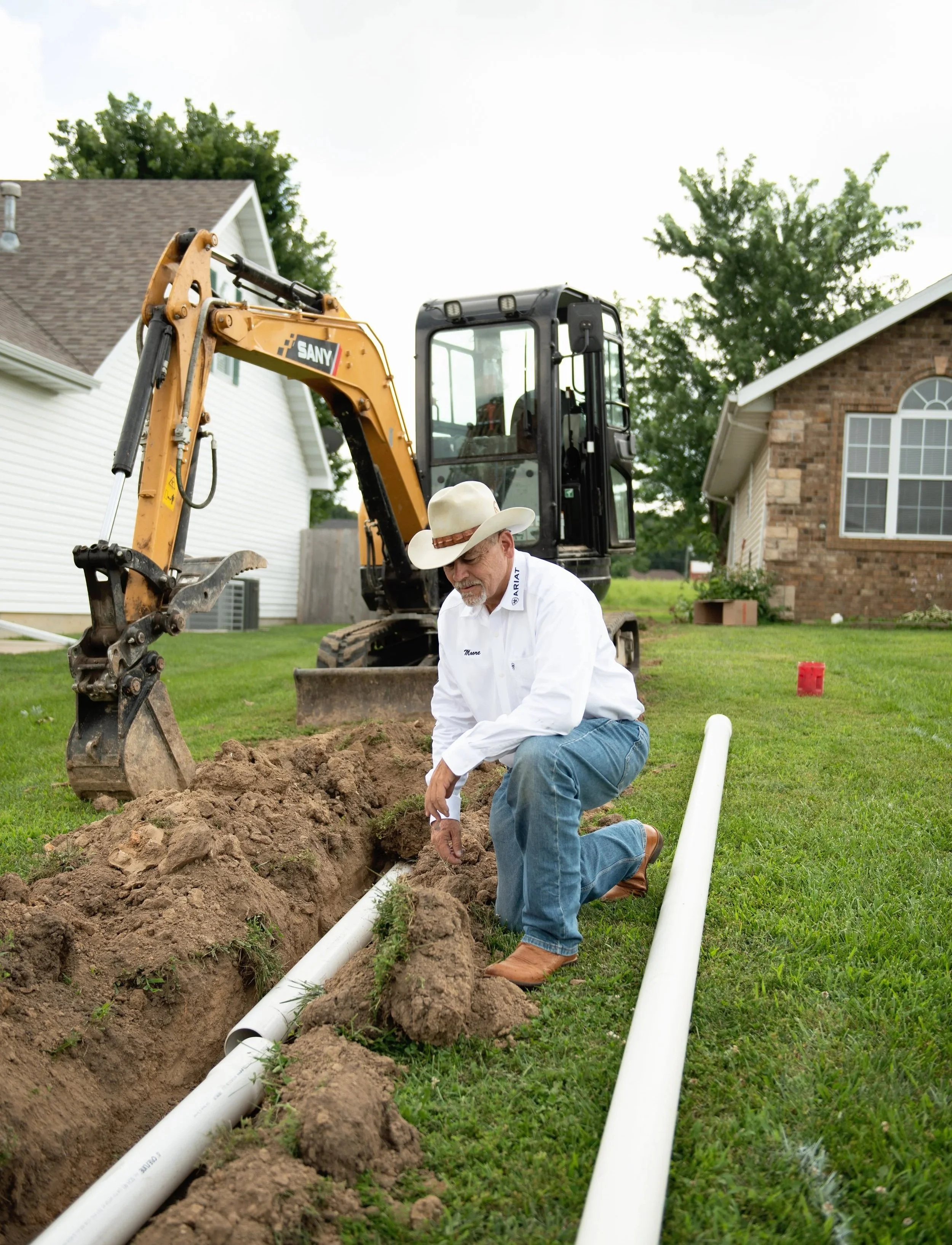 A man in a cowboy hat kneels in a backyard trench, working on underground pipes with a small excavator nearby, houses and trees in the background.