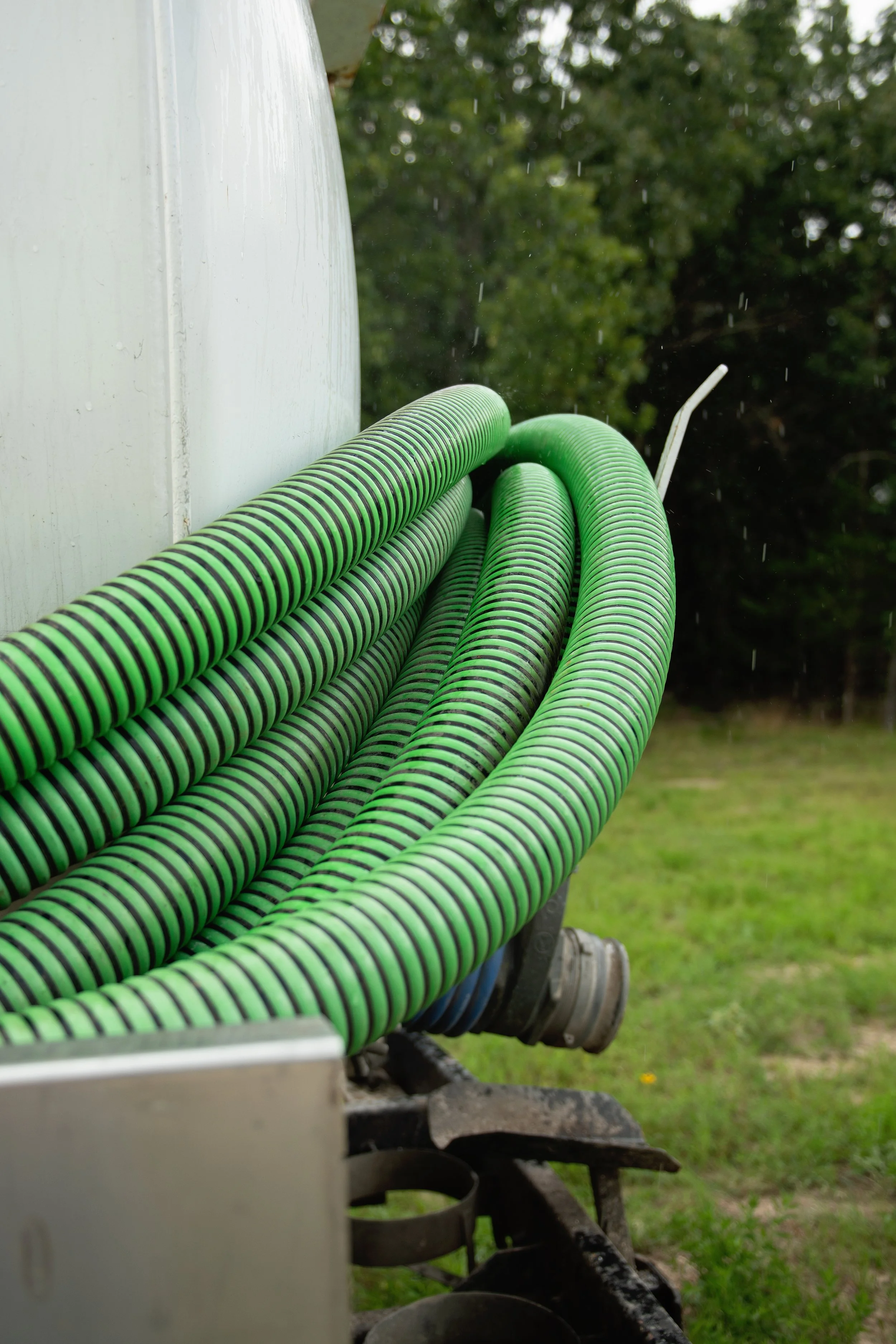 Close-up of green and black striped hoses attached to a piece of machinery outdoors, with trees in the background and rain falling.