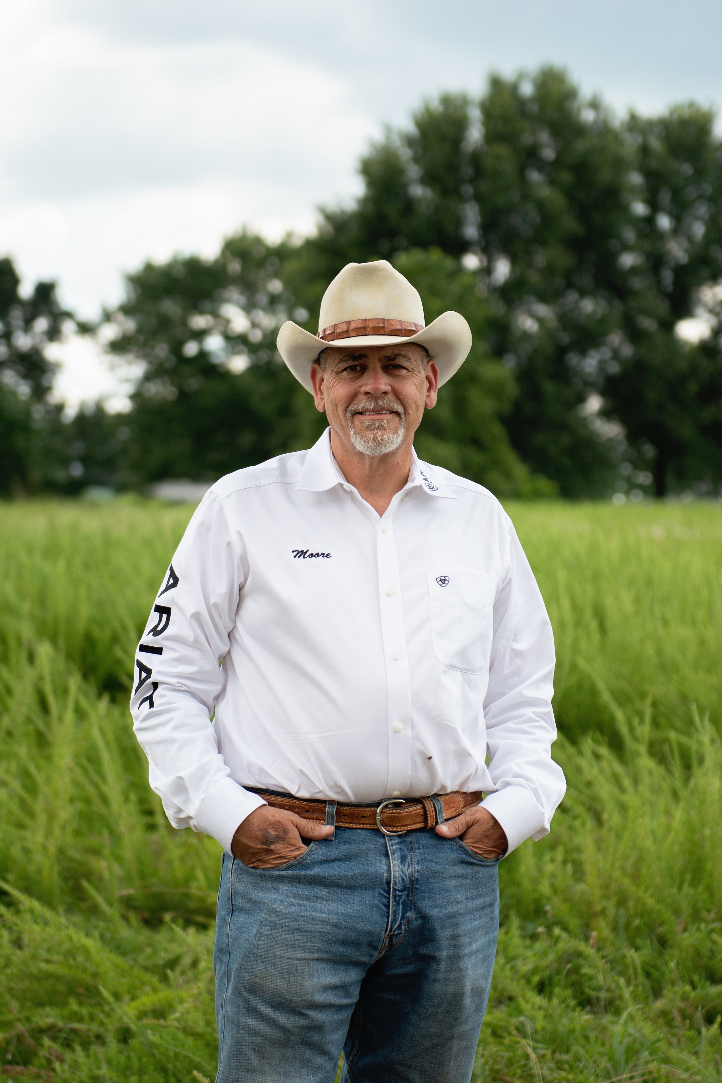 A middle-aged man wearing a cowboy hat and white country shirt with logo and name embroidered on it, standing outdoors in a field with tall grass and trees in the background, smiling at the camera.