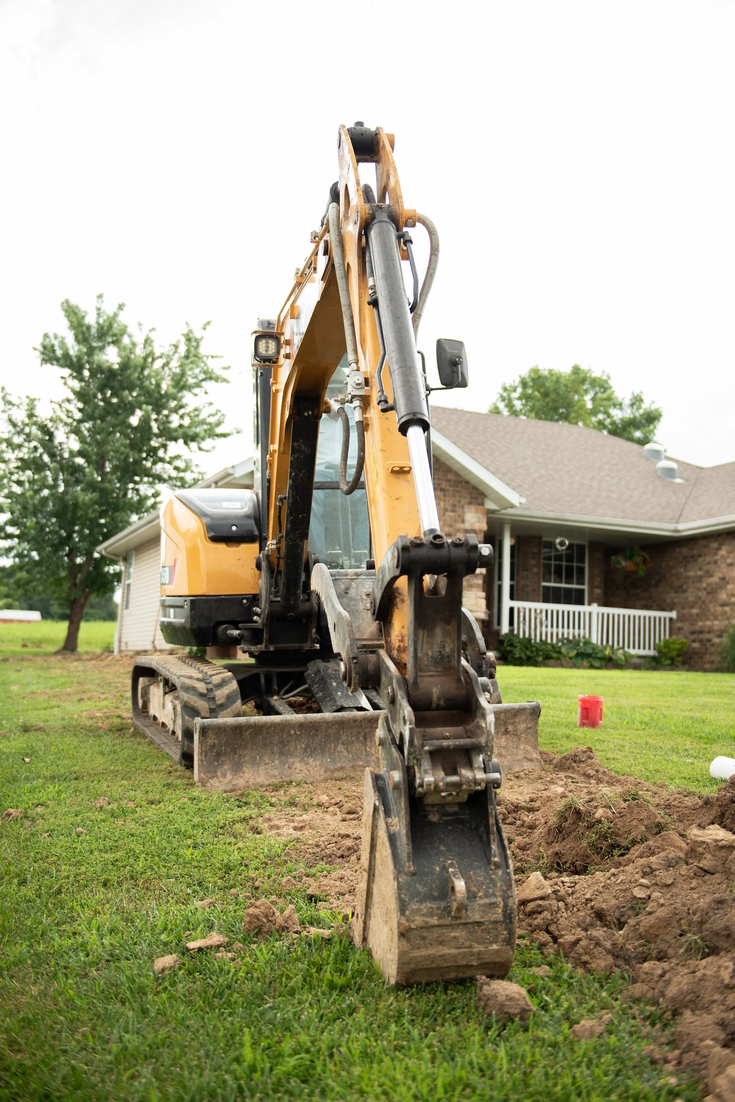 A yellow and black excavator digging a trench in a grassy backyard with a brick house and trees in the background.
