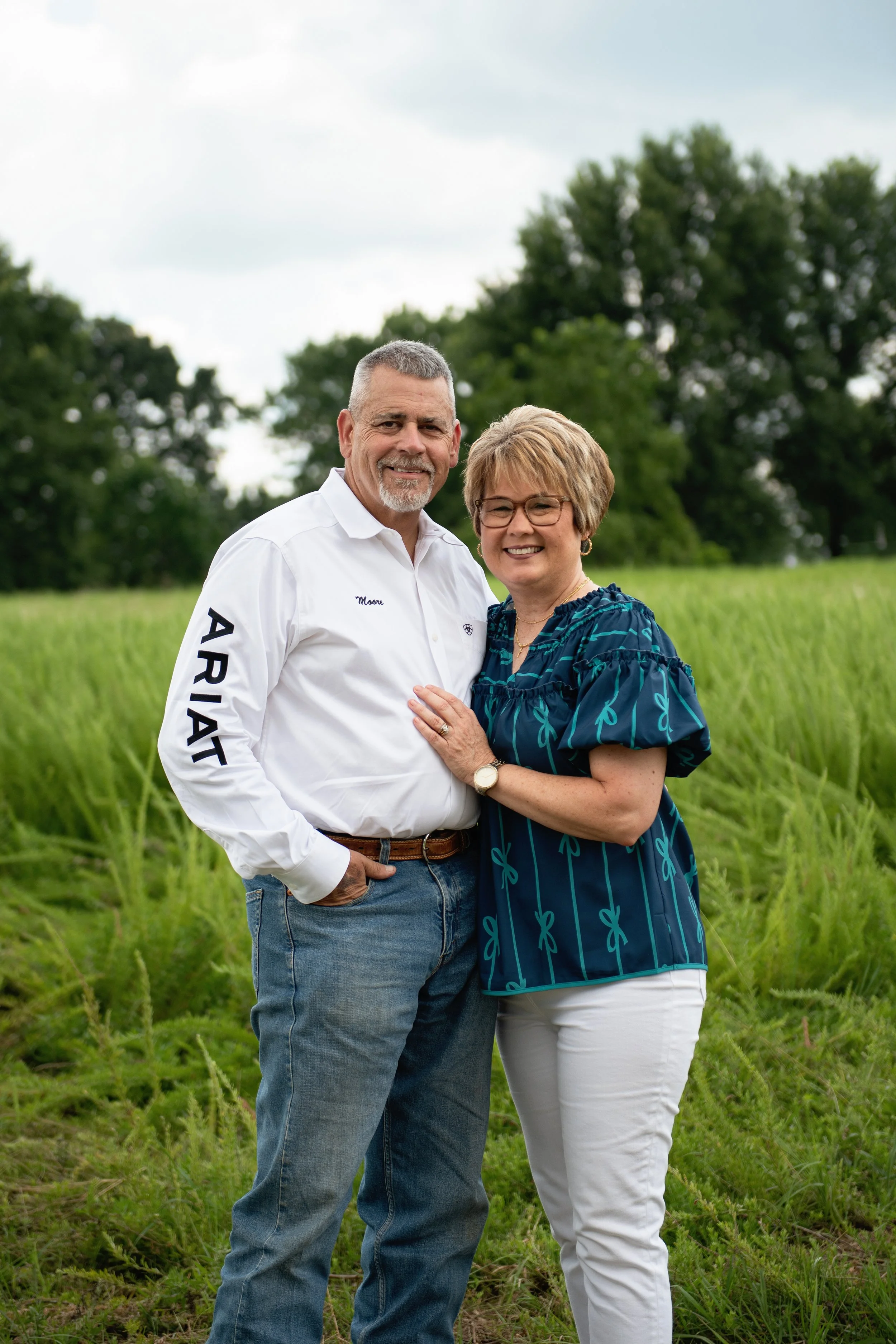 A middle-aged man and woman standing outdoors in a lush green field with trees in the background, smiling at the camera, dressed casually.