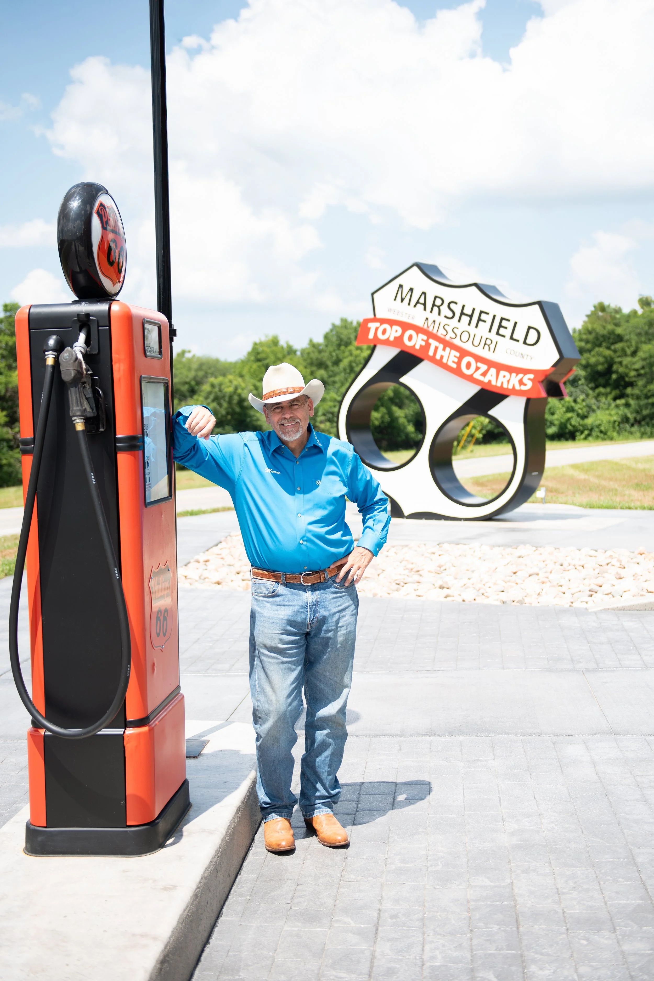 Man in cowboy hat and blue shirt standing next to a vintage orange gas pump with a Route 66 sign and a large roadside sign that says "Marshfield, Missouri, Top of the Ozarks" in the background.