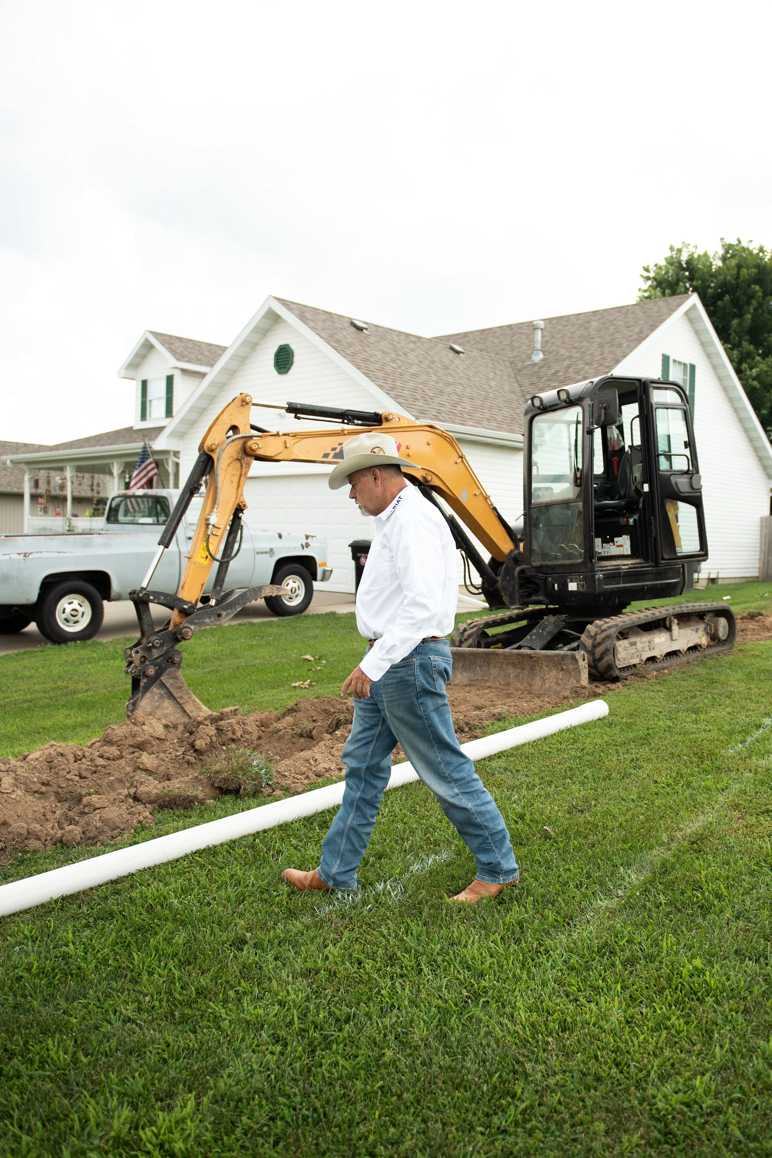 A man in cowboy attire, including a hat and boots, works on installing a pipe in the yard of a suburban house. There is construction equipment and a pickup truck in the background.