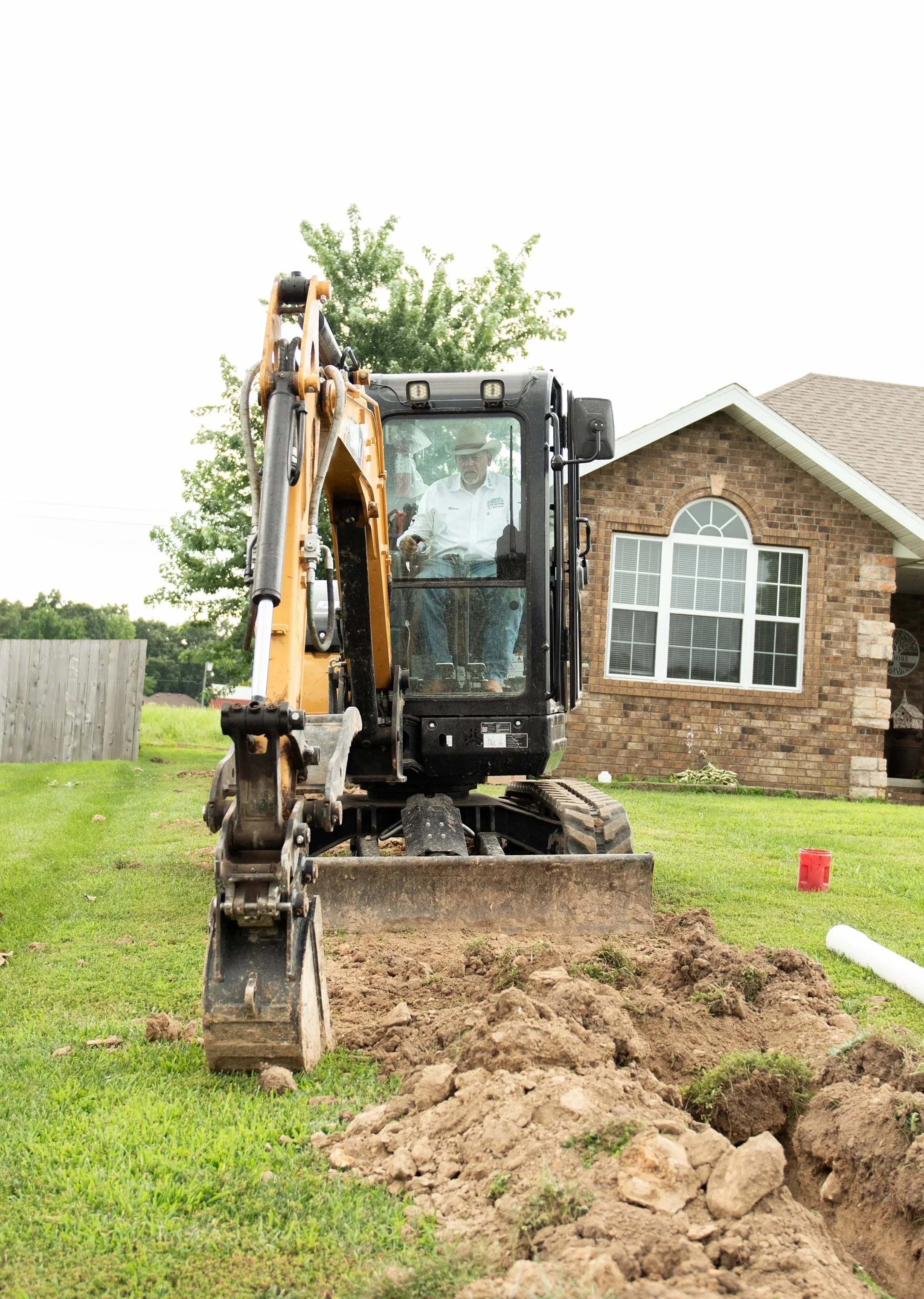 A construction worker operating a small excavator on a residential lawn with a house in the background.