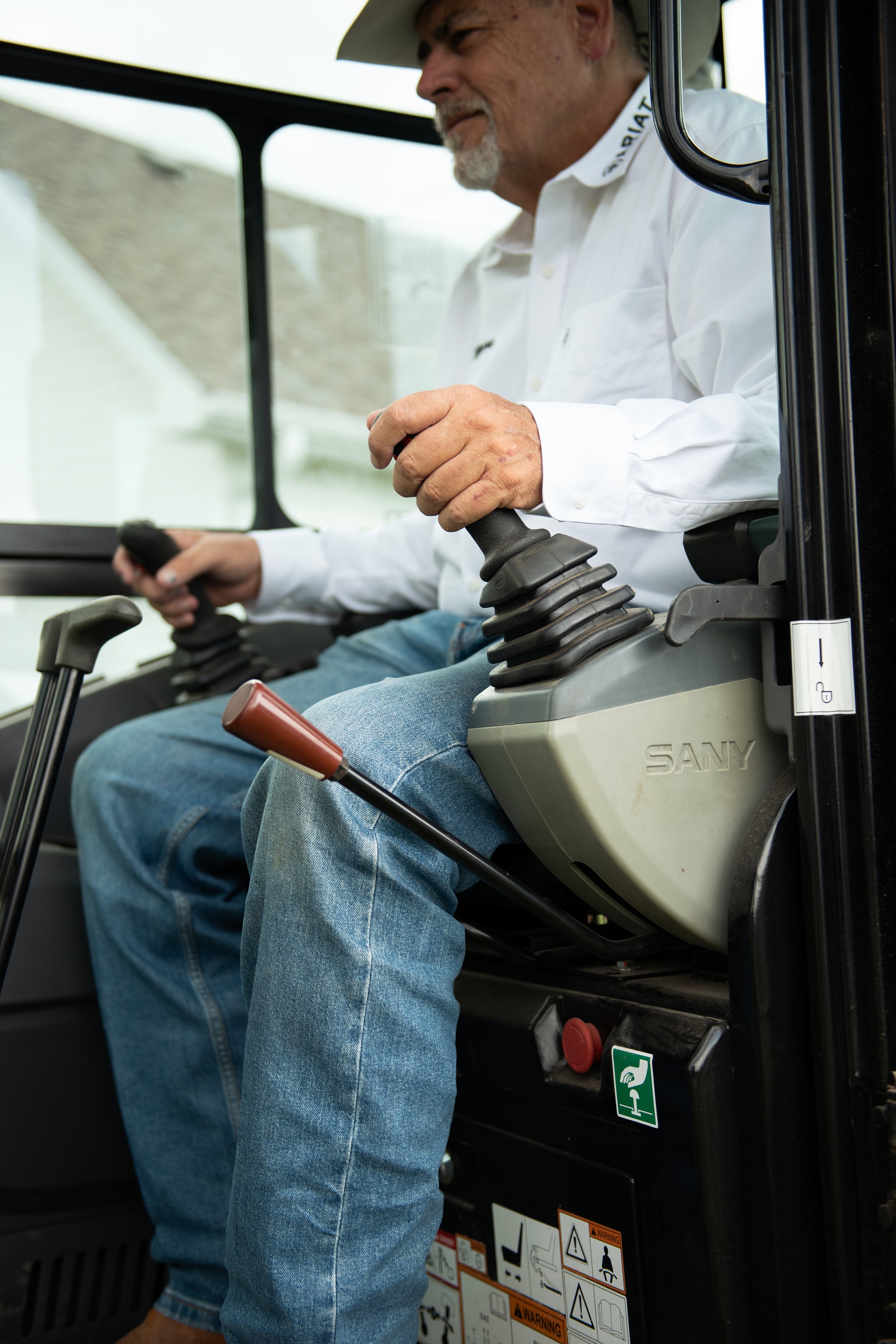 A man with a beard and a cowboy hat operating heavy machinery, possibly a tractor or bulldozer, inside the cab.