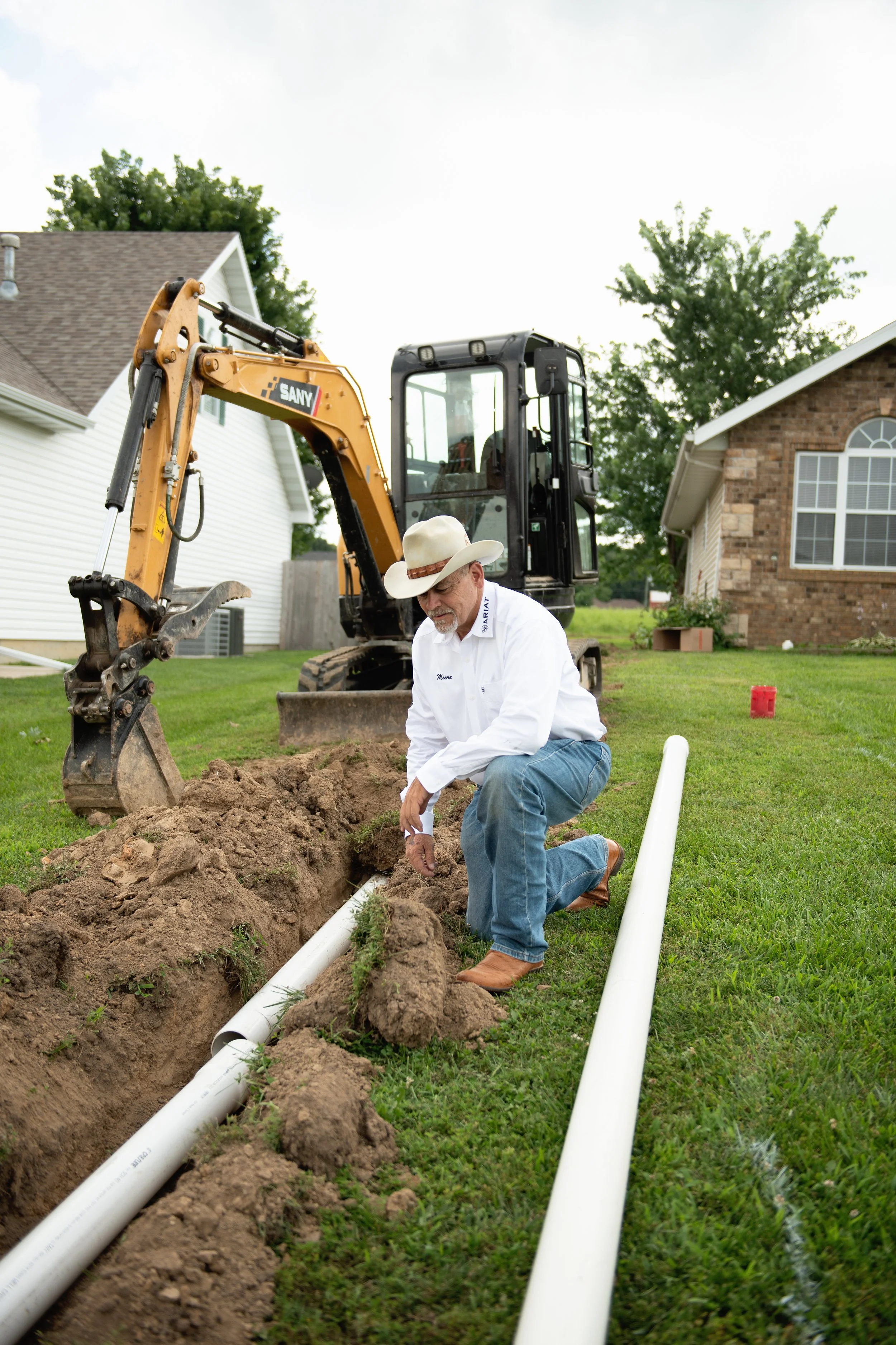 A man in cowboy boots and a white shirt with a hat kneels beside a PVC pipe in a trench in a backyard, with a compact excavator in the background.