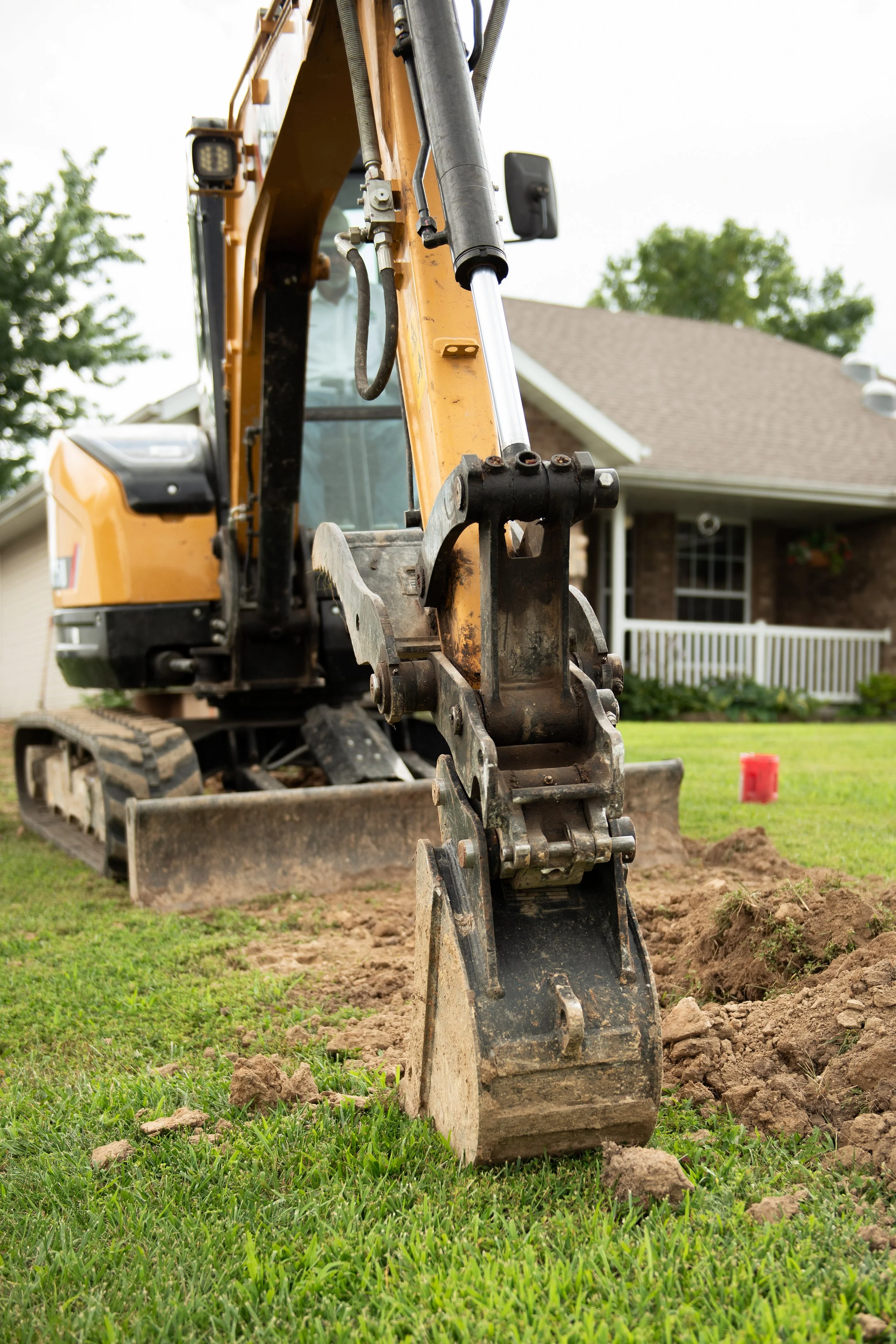 A yellow excavator digging a hole in a grassy yard in front of a house with a white fence and trees.