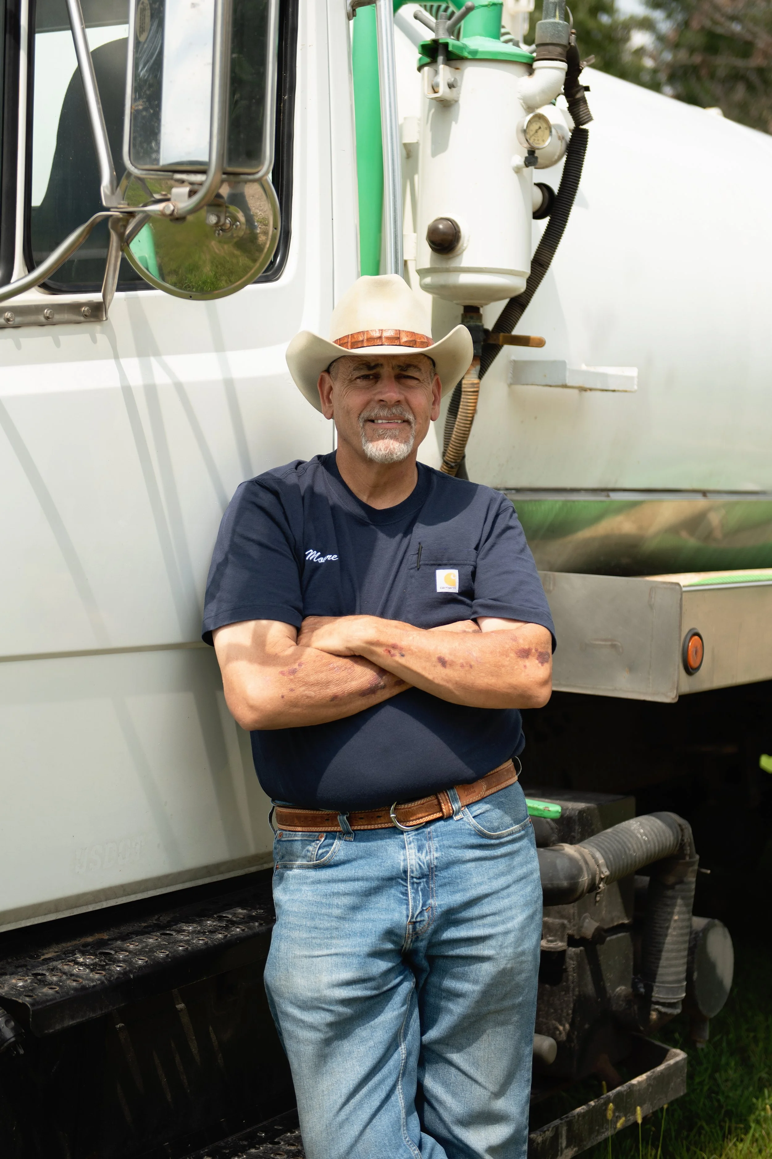 A man with gray hair and beard, wearing a cowboy hat and navy blue t-shirt, standing with arms crossed in front of a white truck with industrial equipment attached.