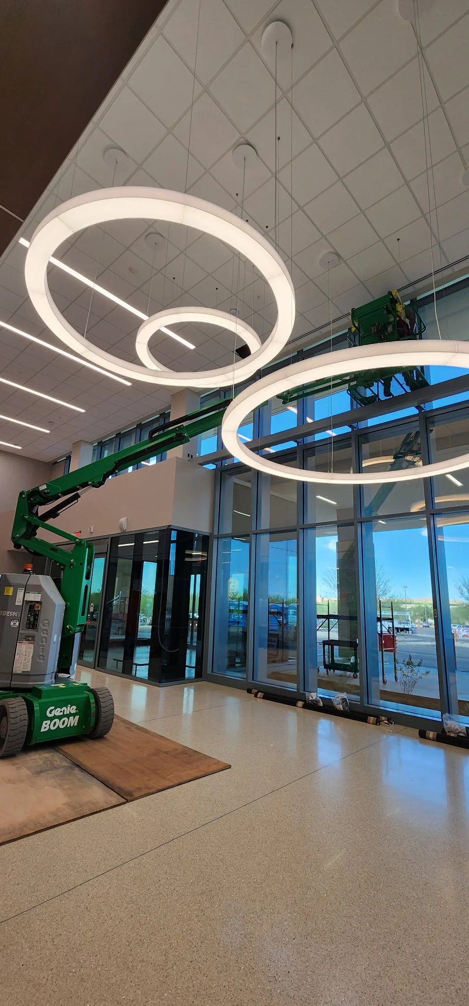 Interior of a building under construction or renovation with large glass windows, modern LED circular light fixtures hanging from the ceiling, and a green scissor lift with a raised platform.