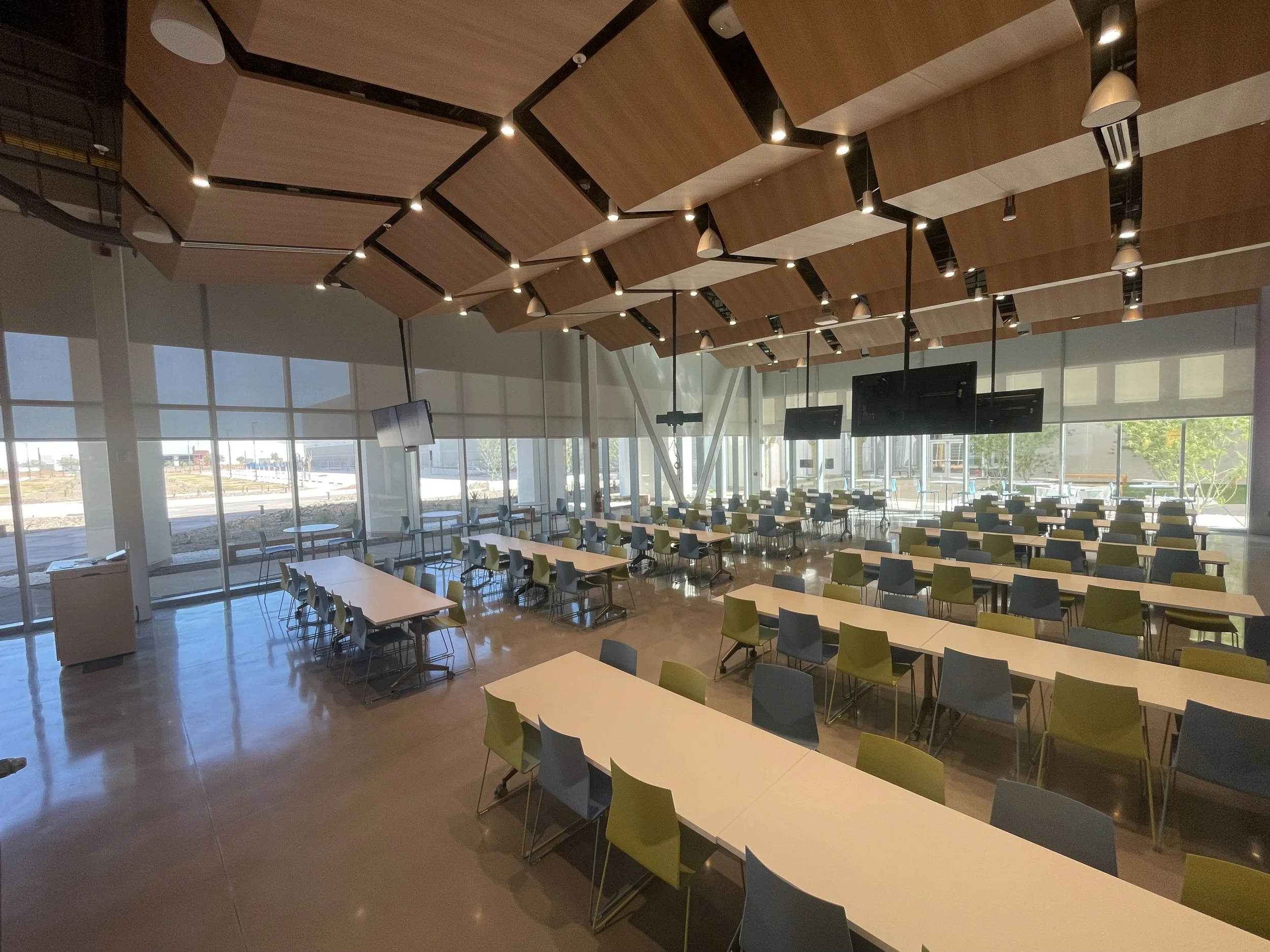 An interior of a modern spacious conference room with a high ceiling featuring geometric wooden panels and hanging lights. The room has large windows allowing natural light and contains multiple tables and chairs arranged for meetings or events.