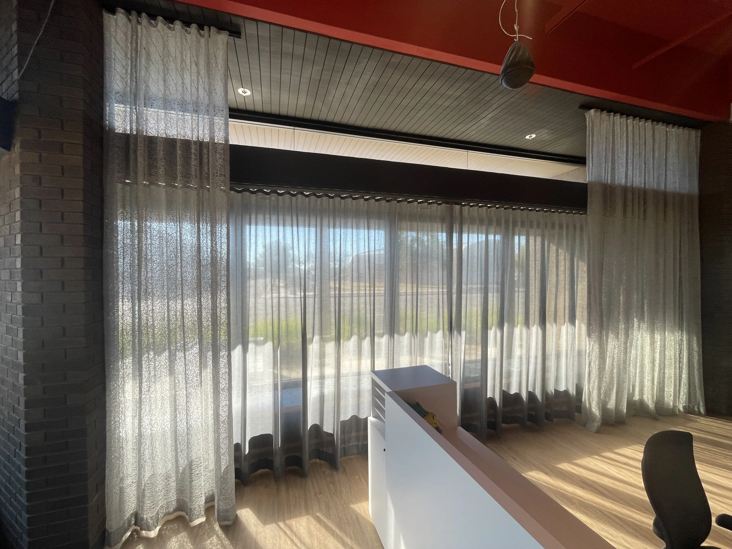 A room with sheer curtains hanging in front of large windows, allowing sunlight to fill the space. The ceiling has wooden paneling painted in a neutral color, and there is a hanging speaker or smoke detector near the ceiling. Part of a white desk or 