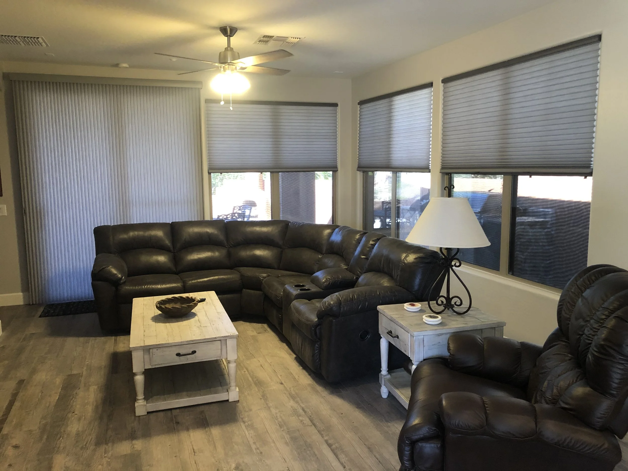 Living room with a dark brown leather sectional sofa, a light-colored wooden coffee table, a side table with a lamp, and several windows with closed blinds. Overhead ceiling fan and light fixtures are also visible.
