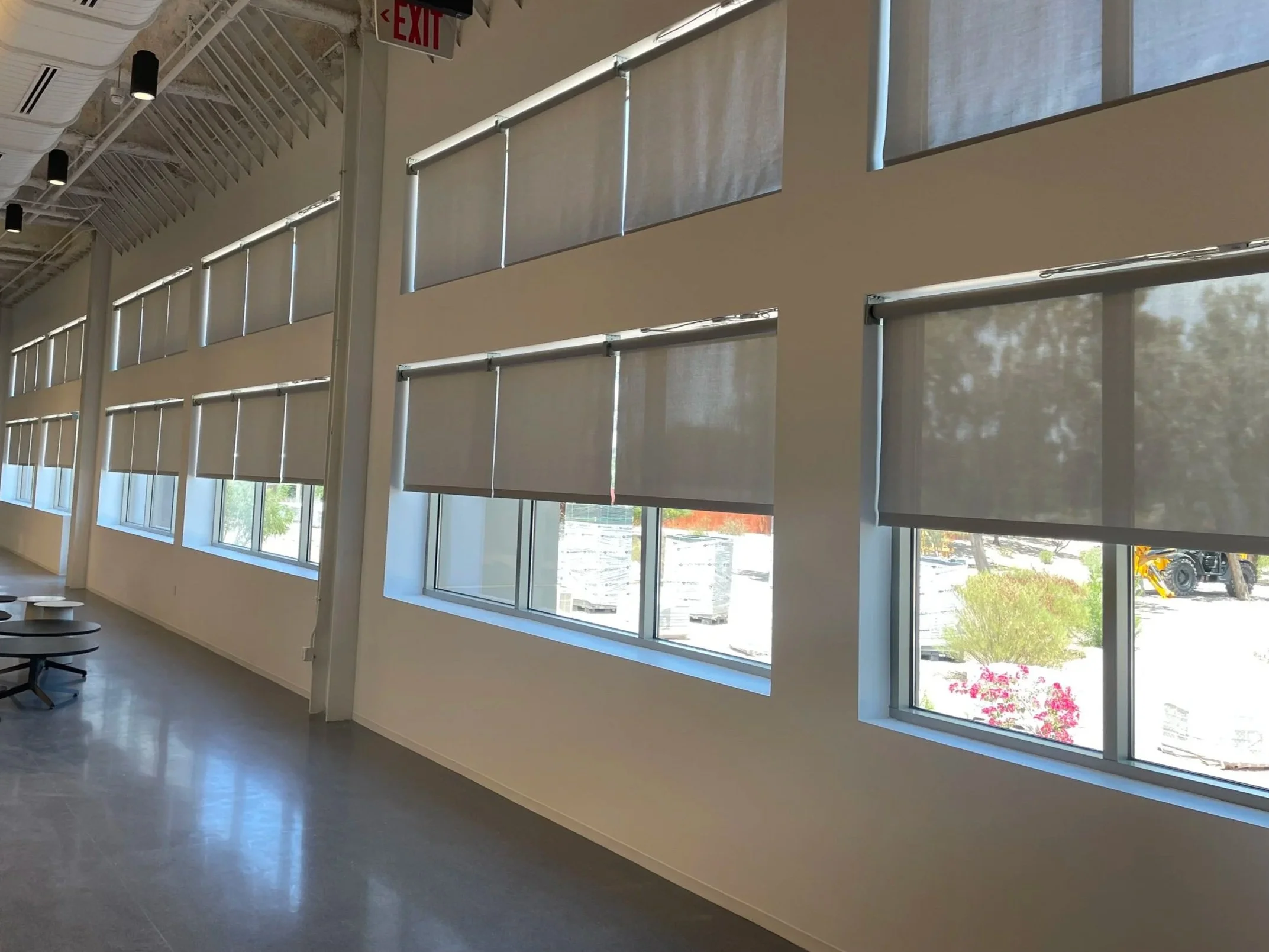 Interior of a modern building with large windows covered by partially drawn roller shades, white walls, and a polished gray floor. There is an exit sign hanging from the ceiling, and part of a staircase with a white railing is visible near the top le