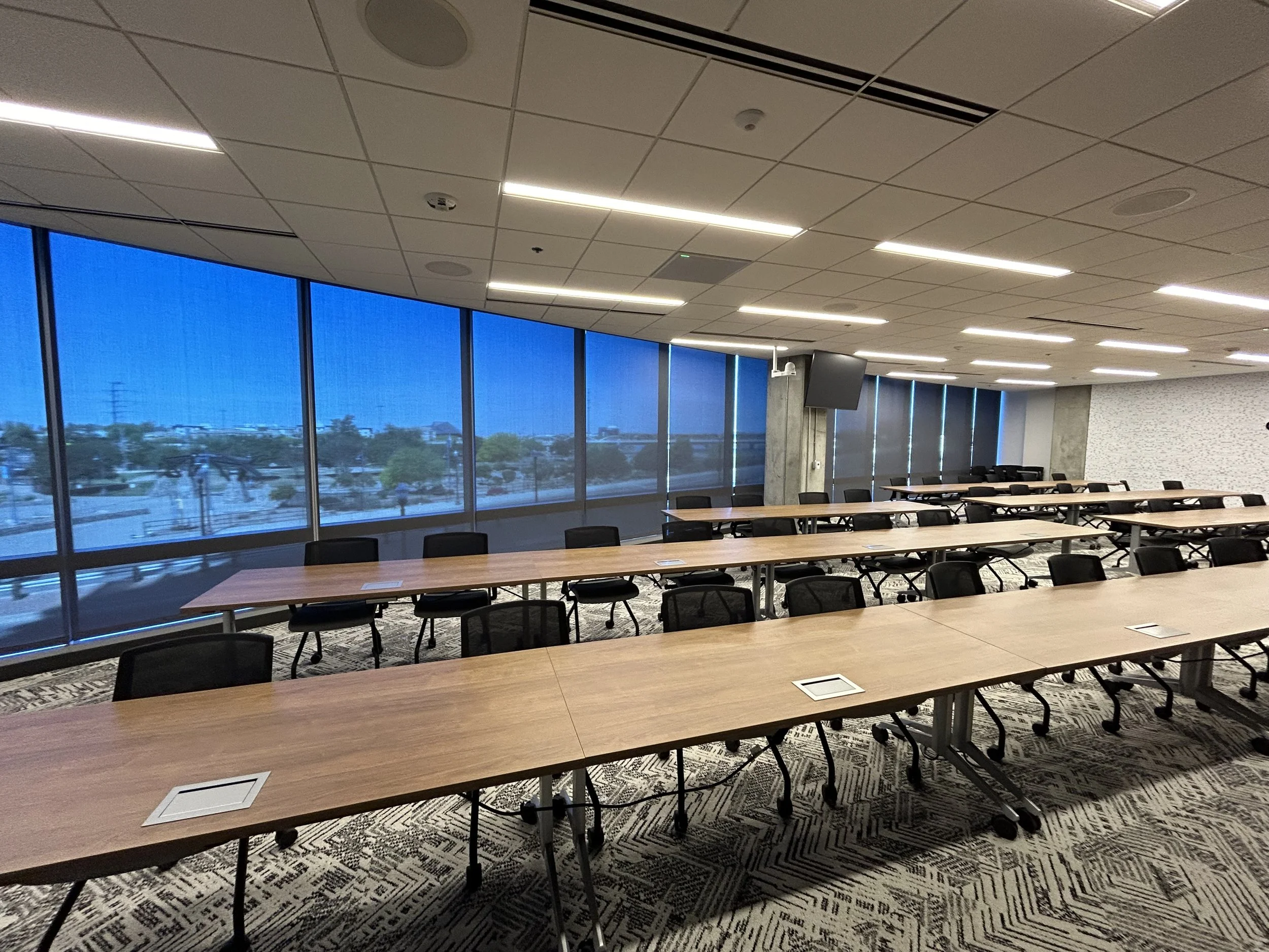 Conference room with long tables, black chairs, large windows showing an outdoor view, and ceiling lights.