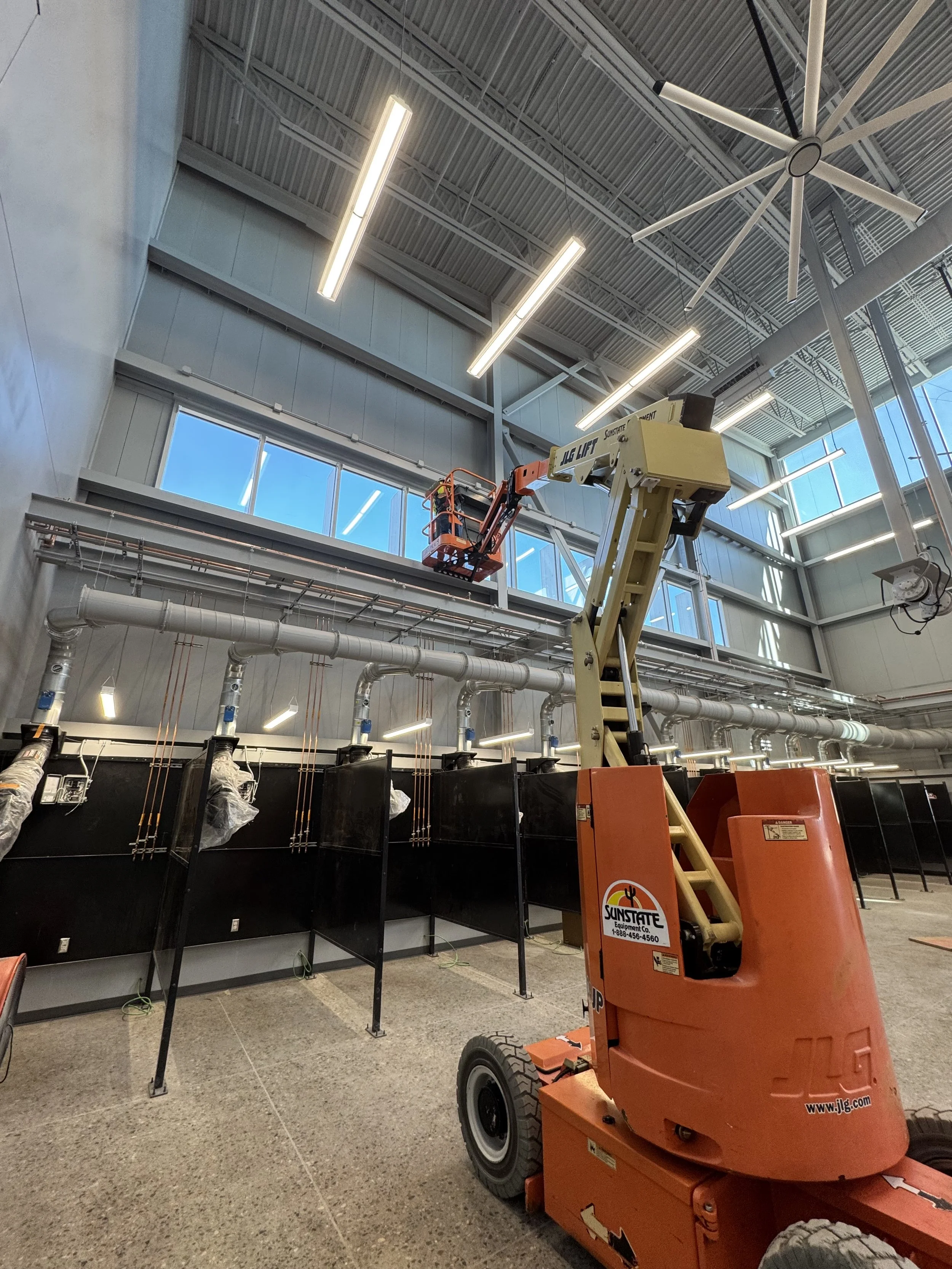 Inside a building with high ceilings, a scissor lift with an orange base is raised, and there are black counters or workstations beneath it. There are large windows and modern lighting fixtures on the ceiling.