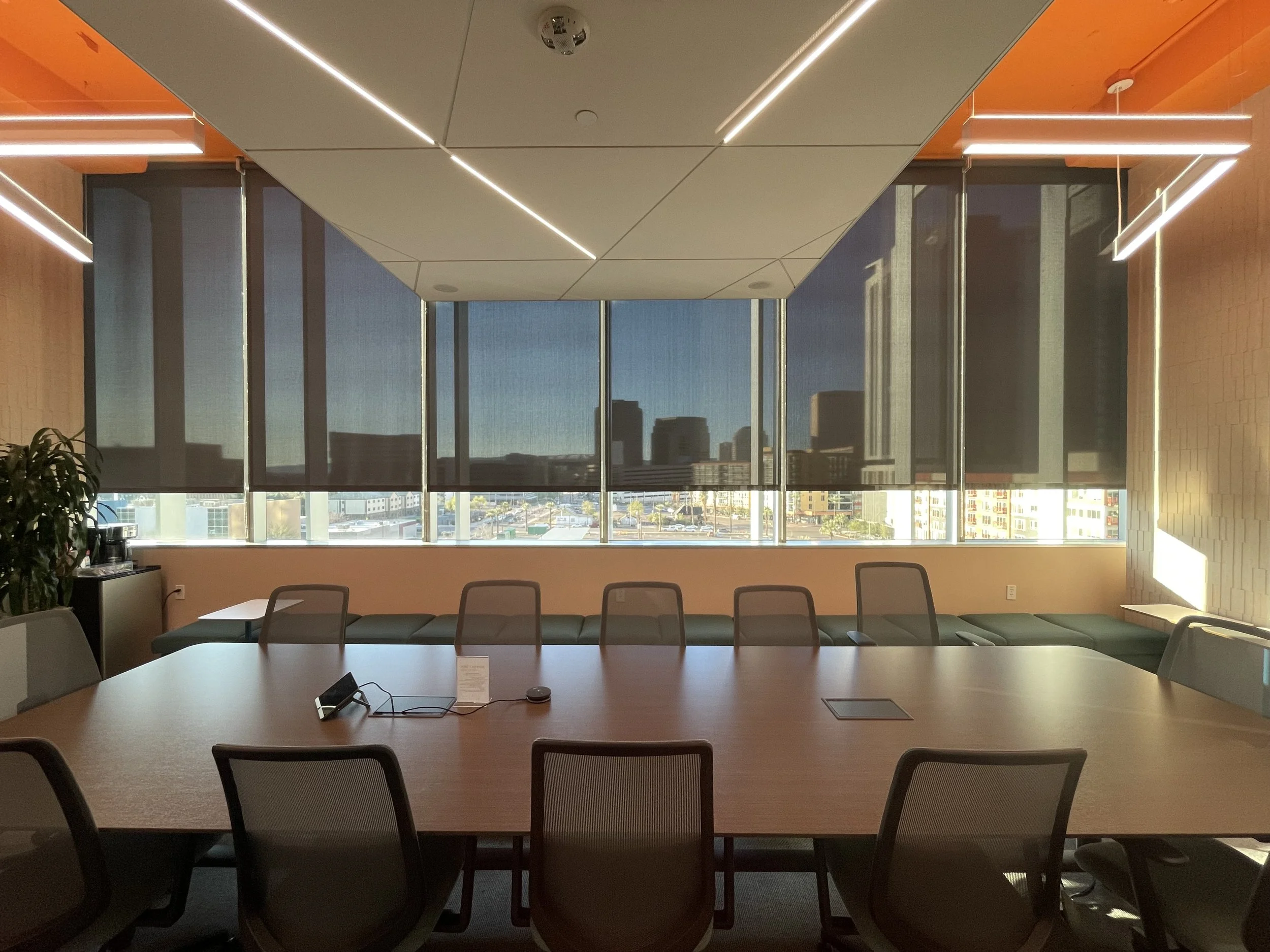 A modern conference room with a large oval table, chairs around it, and large windows with a city view. The ceiling has linear lighting and the room has a warm color scheme with orange and beige tones.