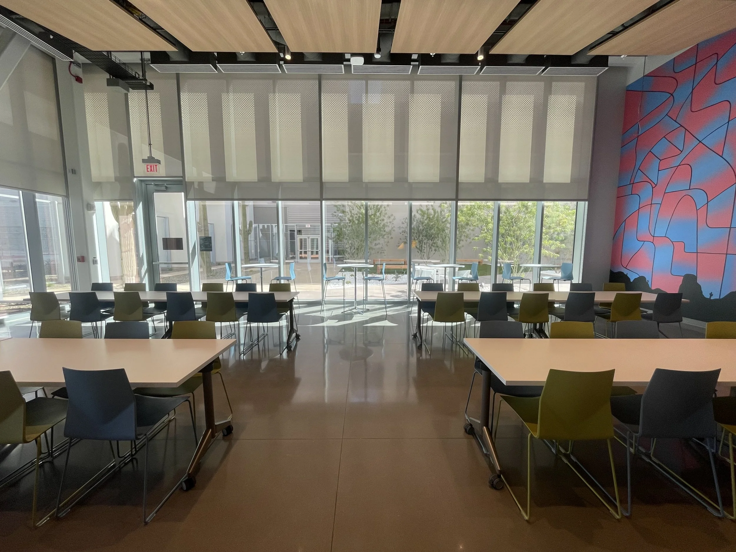Interior of a modern building with large glass windows, tables, and chairs, and a colorful geometric mural on the wall.