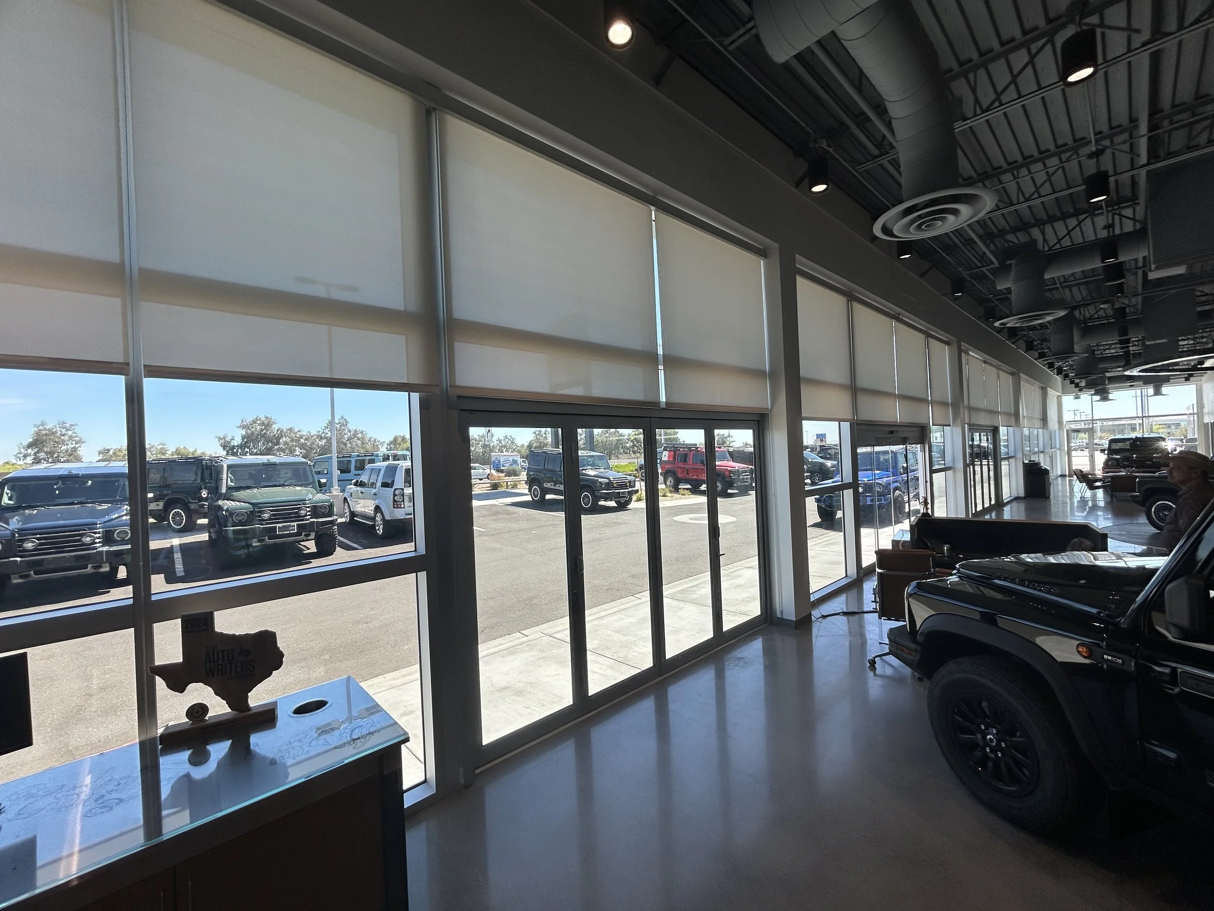 View of a car dealership with large glass windows revealing parked cars outside and some cars visible inside the showroom. The ceiling features black industrial-style ductwork and circular air vents with lighting fixtures.