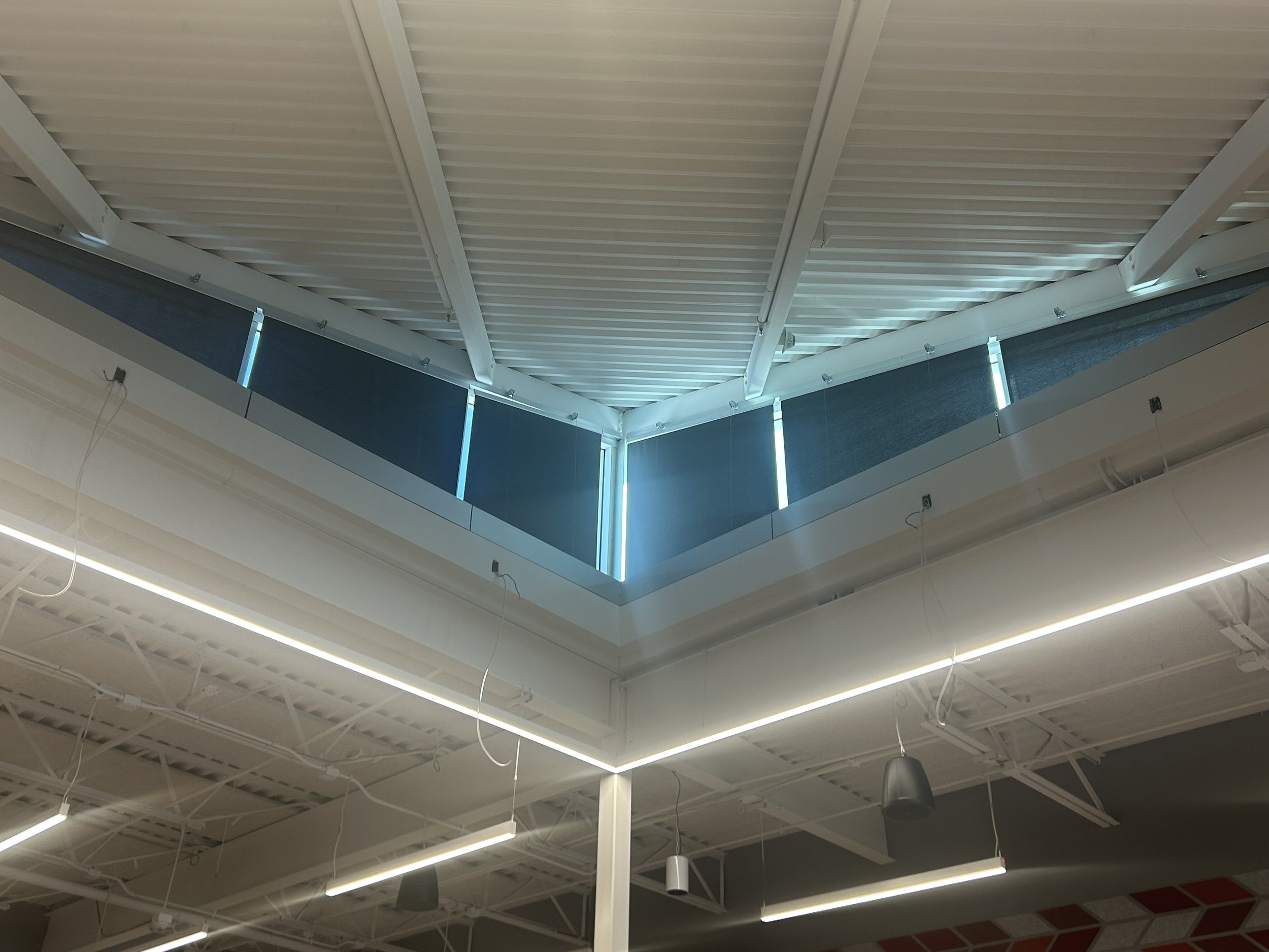 The ceiling of a modern building with white beams, corrugated panels, LED strip lighting, and hanging black speaker and light fixtures.