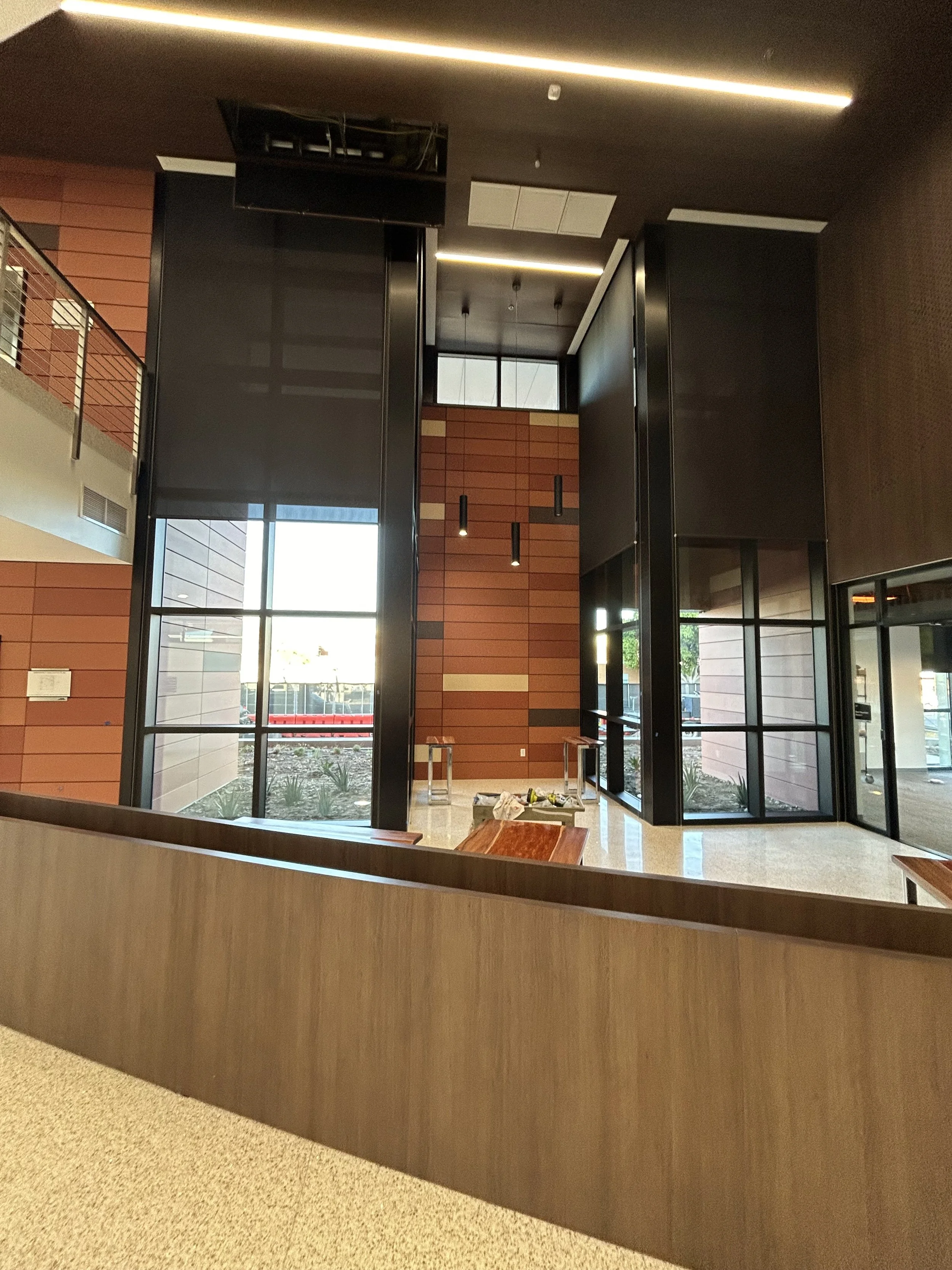 Interior of a modern commercial building with large windows, wooden wall accents, and a high ceiling with recessed lighting. Construction materials and tools are on the floor.