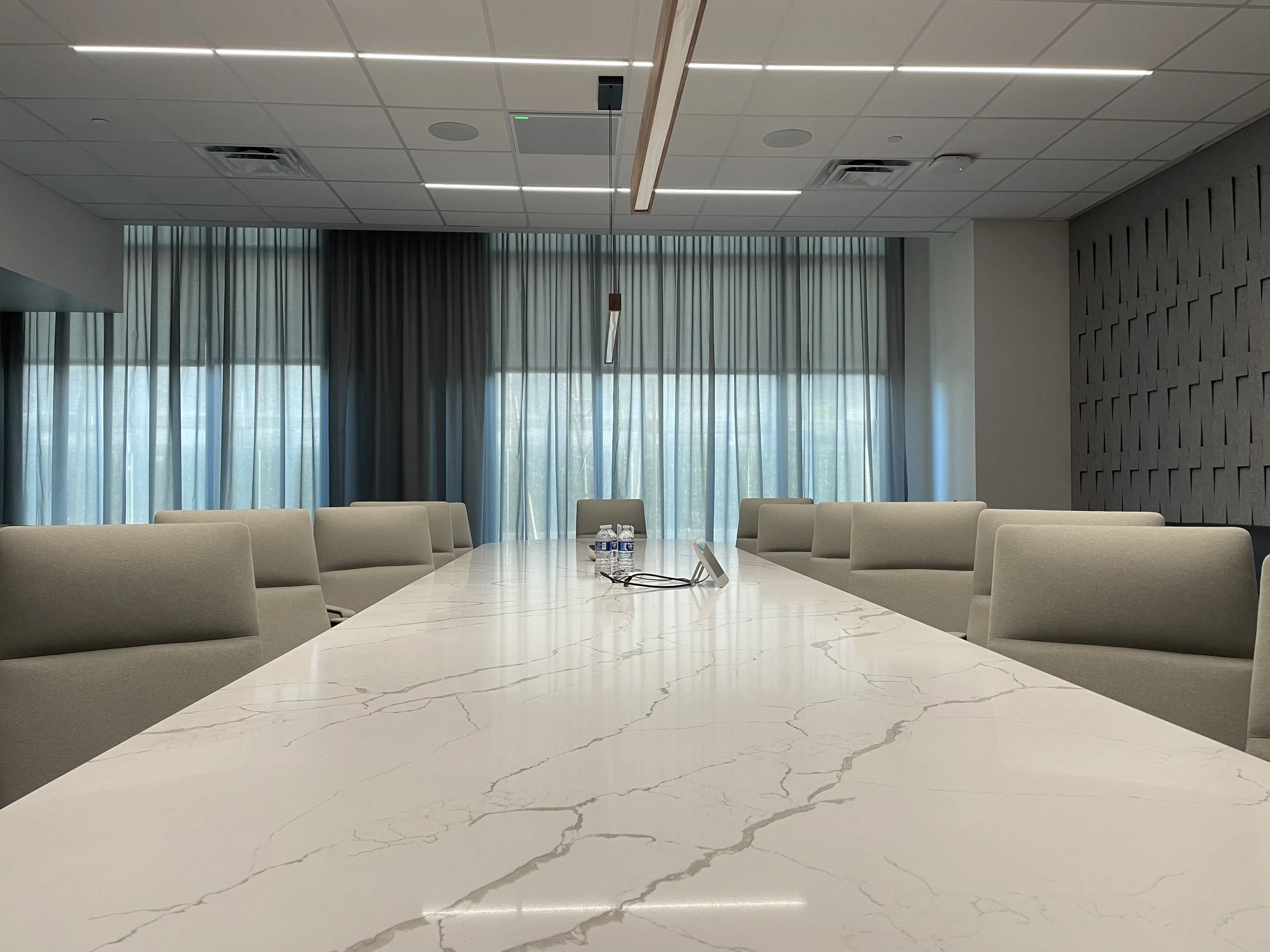 A conference room with a large white marble table, surrounded by beige chairs, with a row of water bottles and conference materials in the center. The back wall features large windows with curtains, and the ceiling has built-in lighting and ceiling v