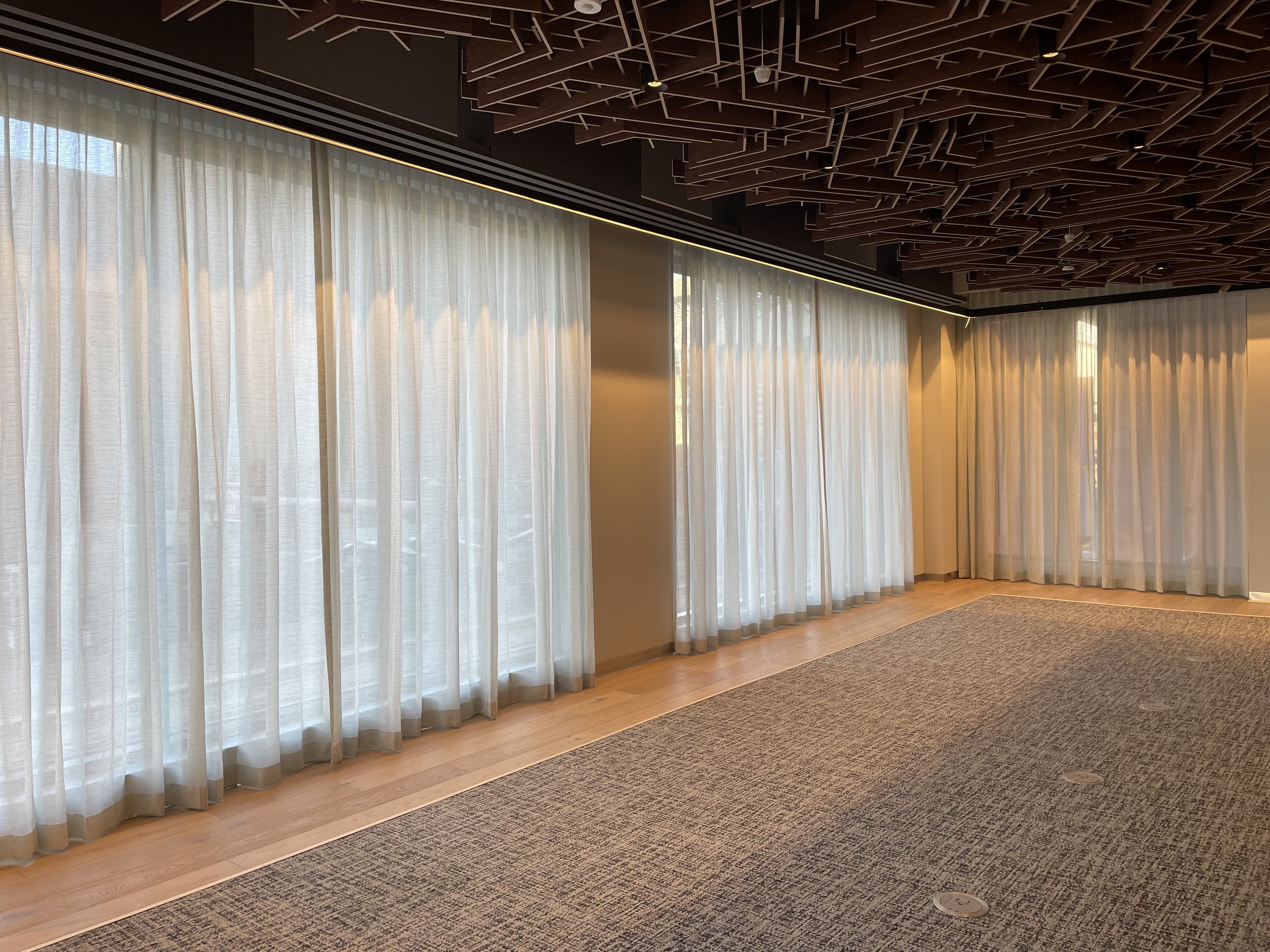 An interior room with large white curtains covering windows, wooden flooring, a gray carpet, and a decorative dark ceiling with a geometric pattern.