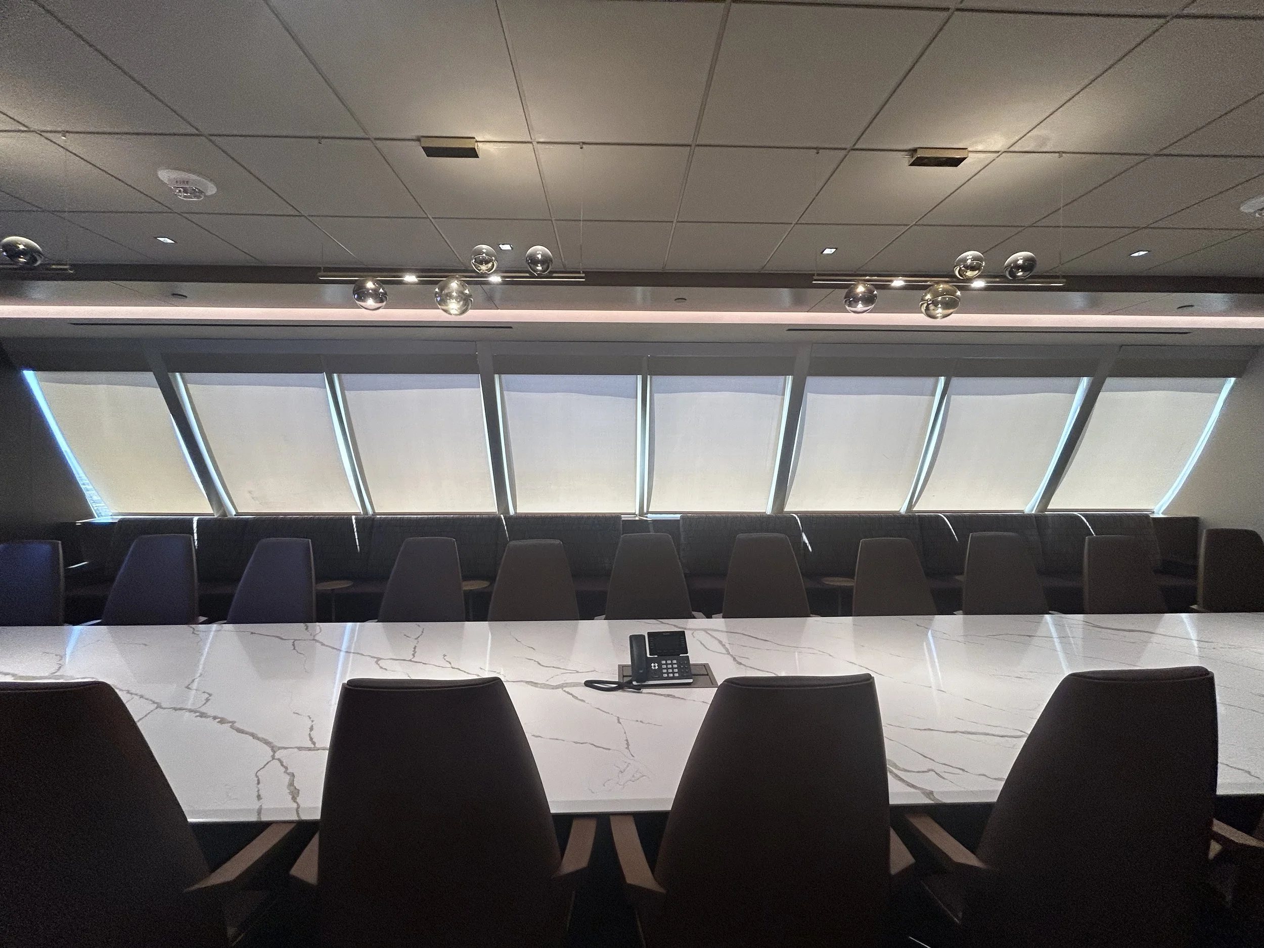 Modern conference room with a long white marble table, black chairs, large windows with blinds, and a phone on the table.