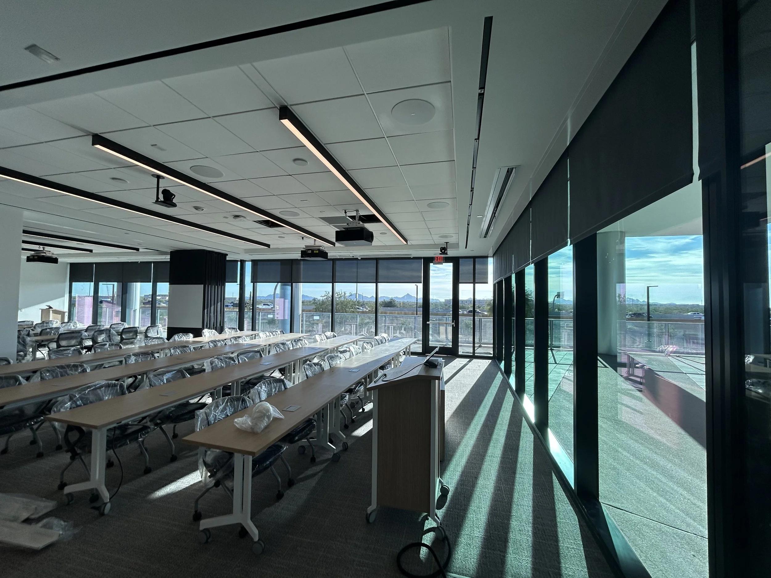 Empty modern conference room with large windows, sunlight, and city view, with rows of chairs and tables covered in plastic.