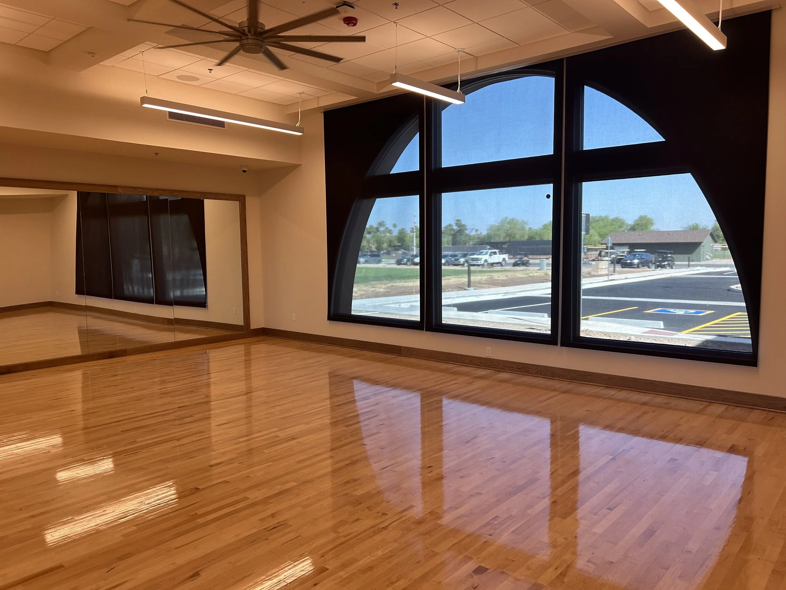 The image shows an empty room with a wooden floor, large black-framed windows, and ceiling lights. Outside the windows, a parking lot and some trees are visible.