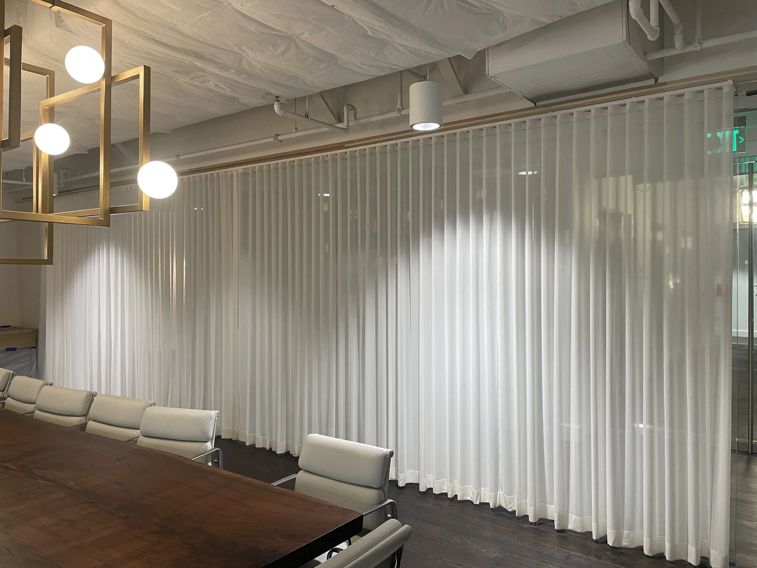 Modern conference room with a long wooden table, white cushioned chairs, white vertical blinds, and a decorative gold geometric chandelier with spherical lights.