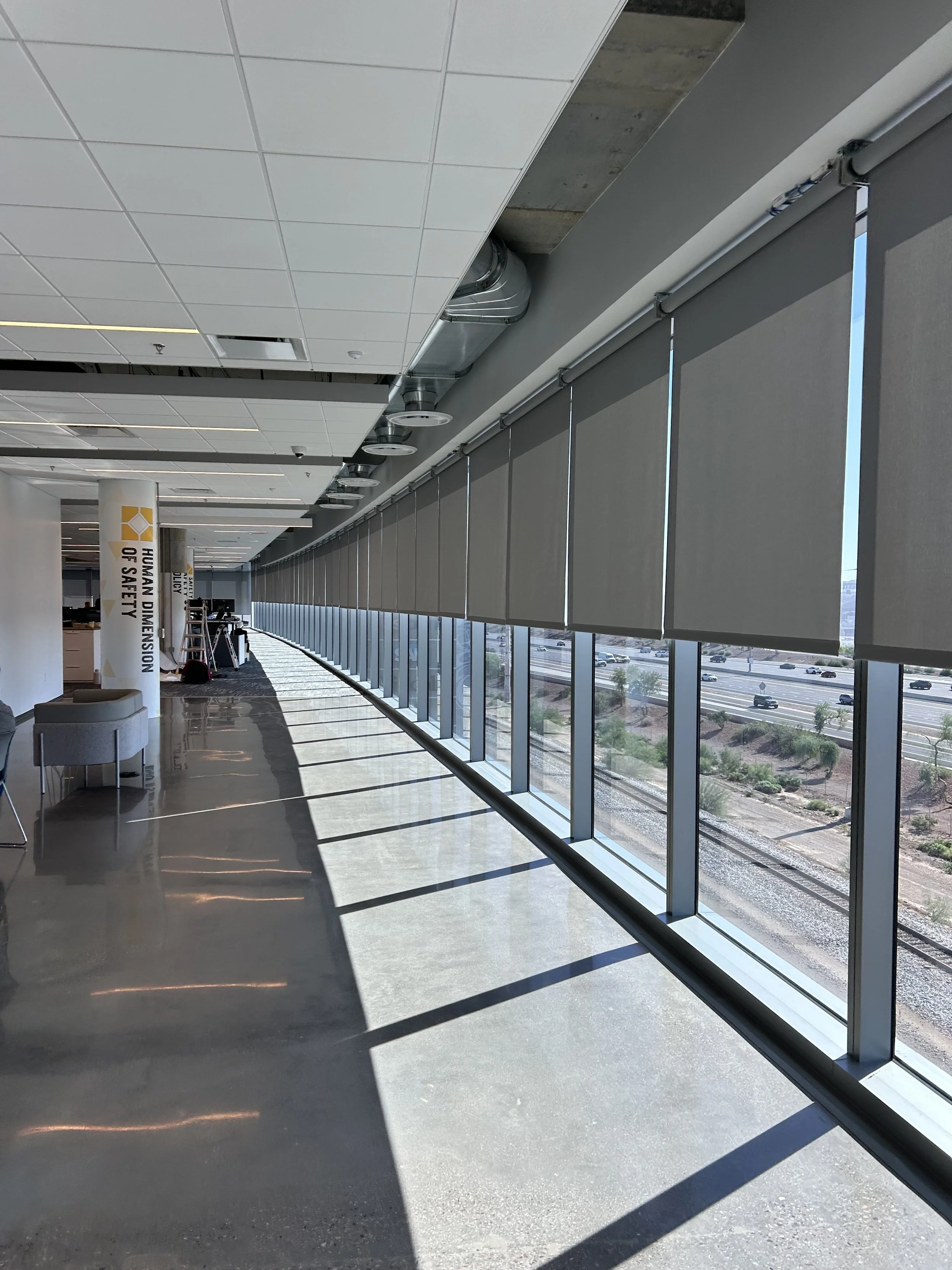 Interior of an airport or office with large windows and closed roller shades, sunlight casts shadows on the floor, modern ceiling with ventilation ducts, a white pillar with safety signage, and some chairs and equipment in the background.