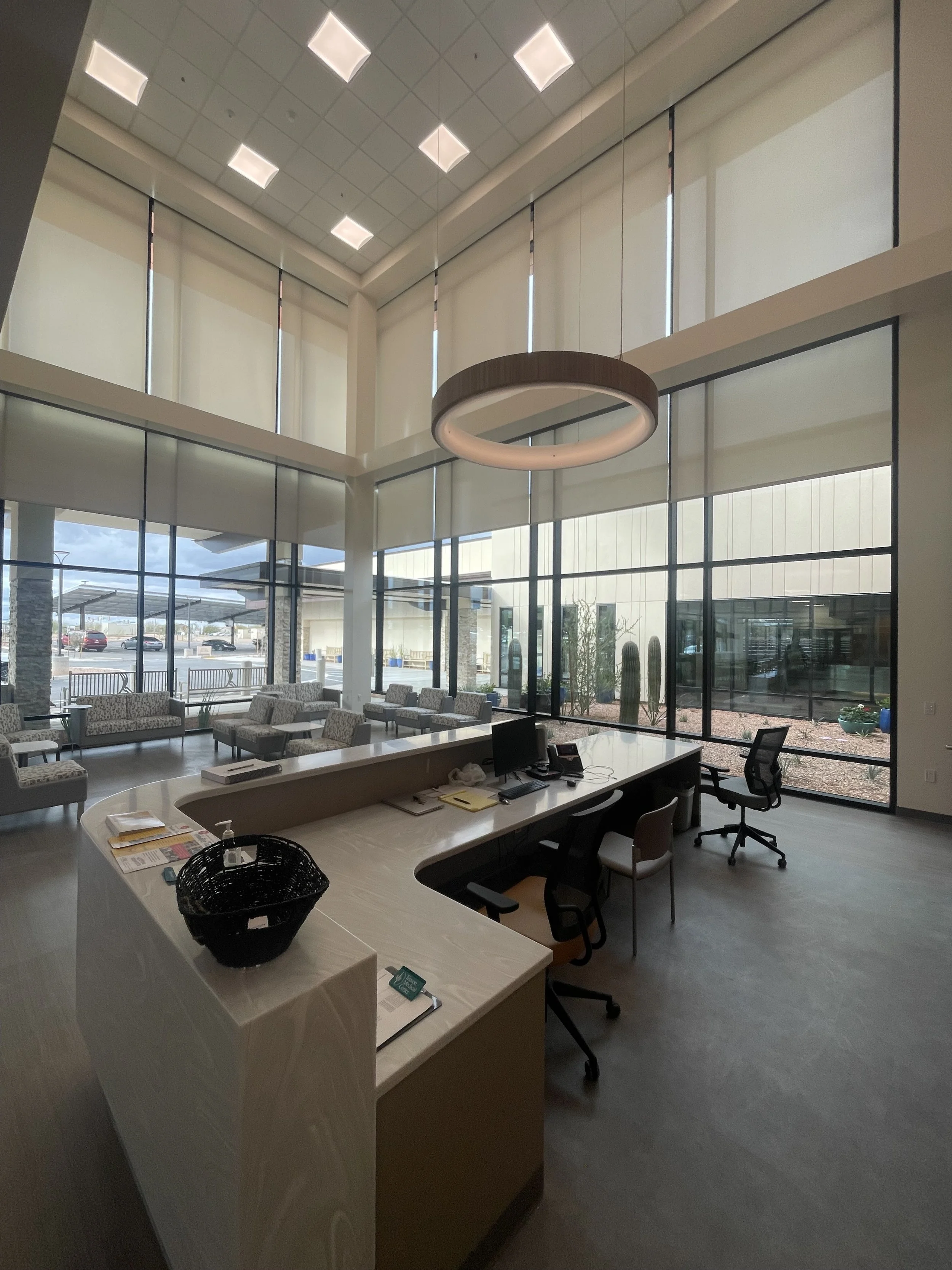 Modern office lobby with large glass windows, seating area with patterned chairs, circular hanging light fixture, and a reception desk with office chairs.