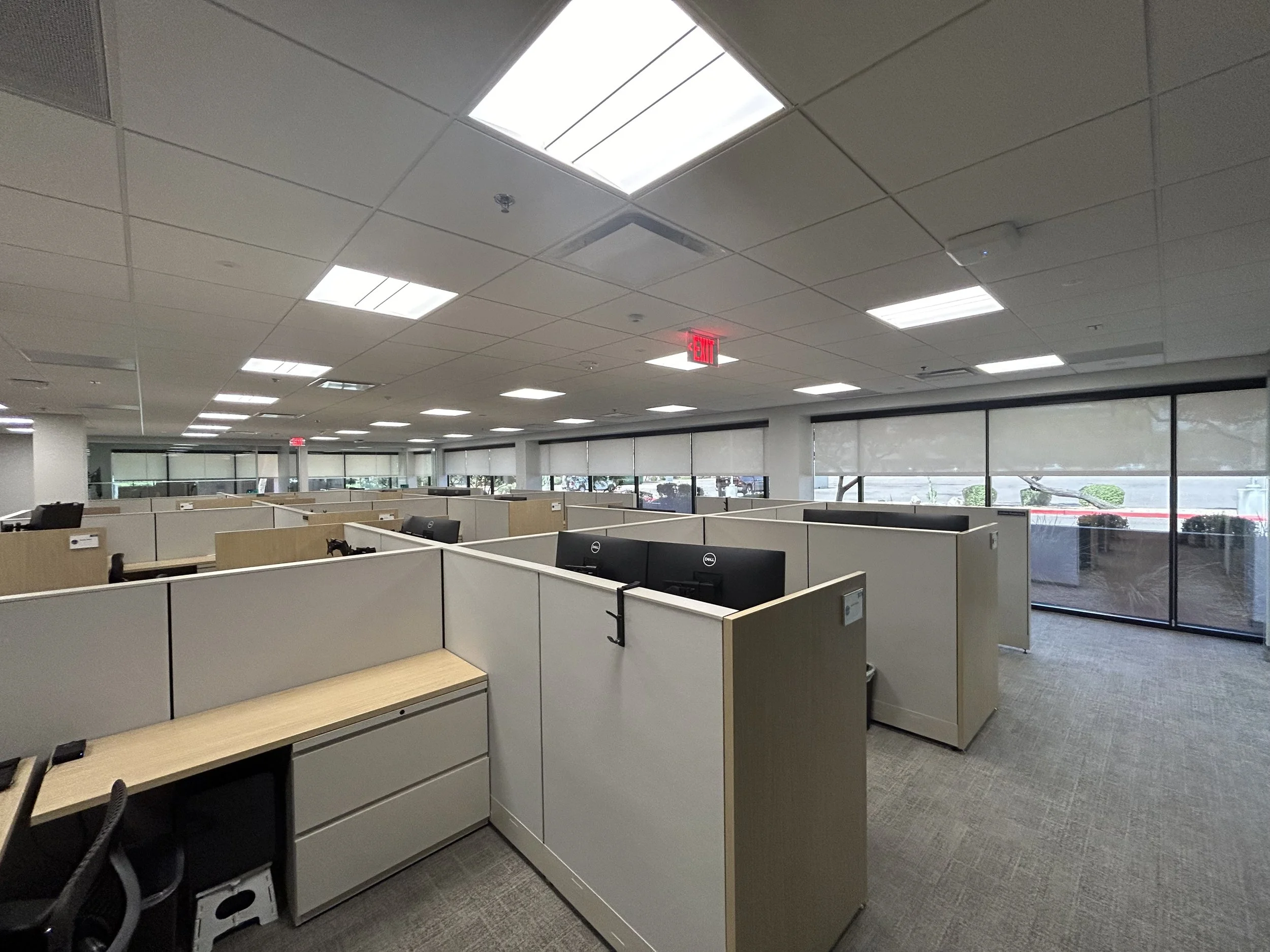 Office cubicles with computers inside a modern office space with large windows and a dropped ceiling with fluorescent lighting.