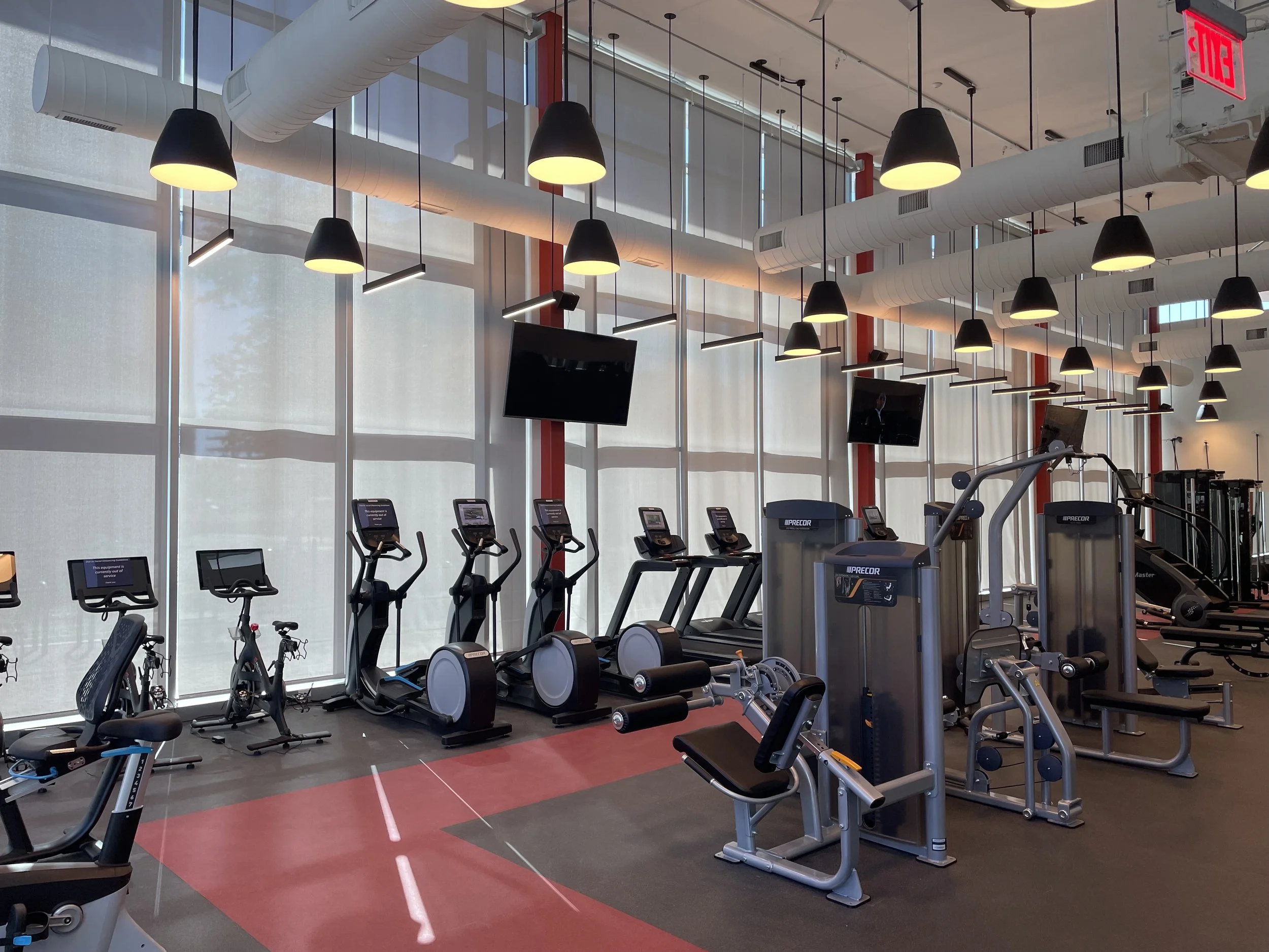 Inside a modern gym with workout equipment such as treadmills and exercise bikes arranged in rows, large windows with blinds, hanging pendant lights, and screens on some equipment.