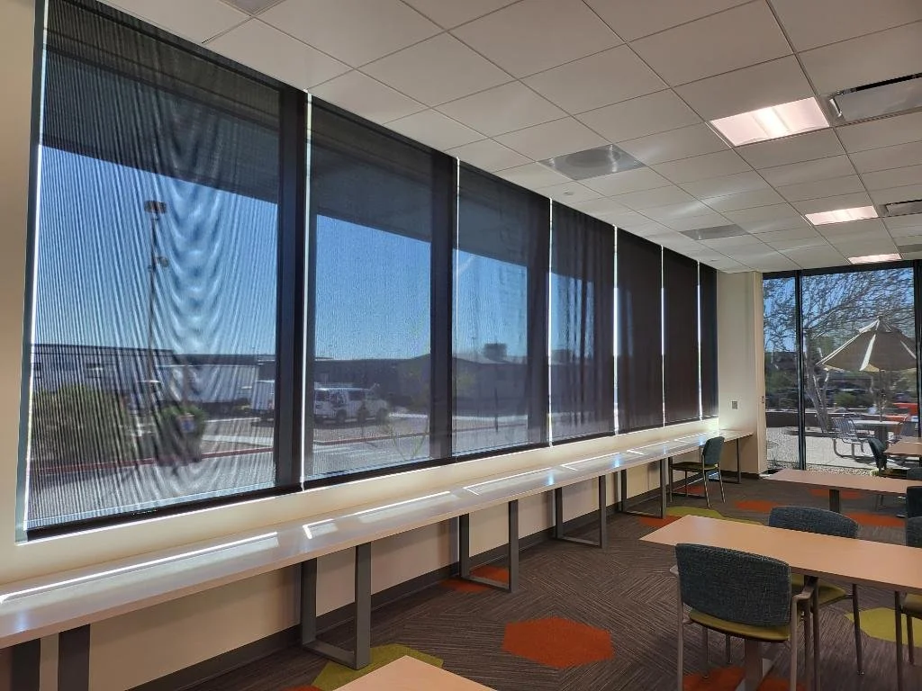 Room with large windows covered by black window shades, some light coming through, with tables and chairs inside, and outdoor patio furniture visible through the window.
